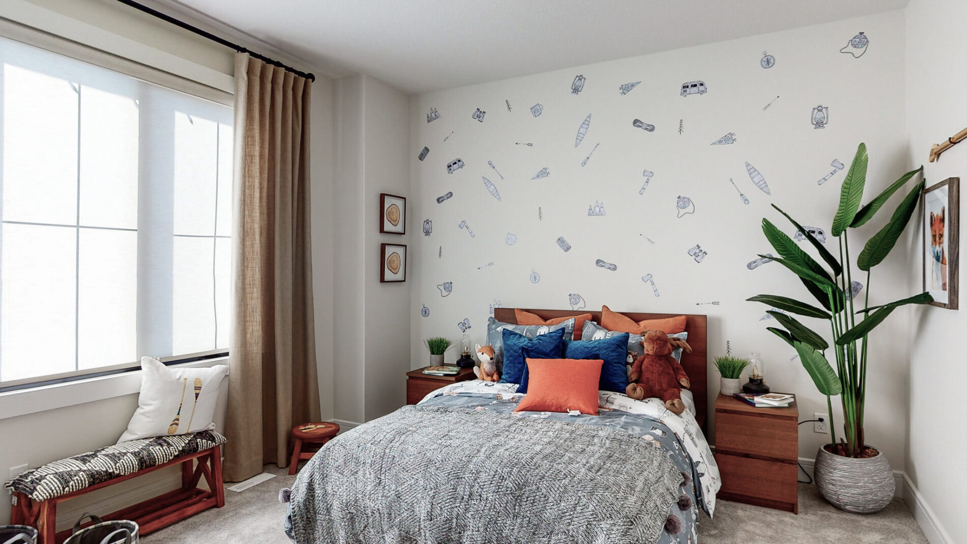 A fun secondary bedroom in the Kalliope showhome with decals on the wall behind the warm wood bed and matching night tables, a large plant in the corner and large black framed windows on the opposite wall