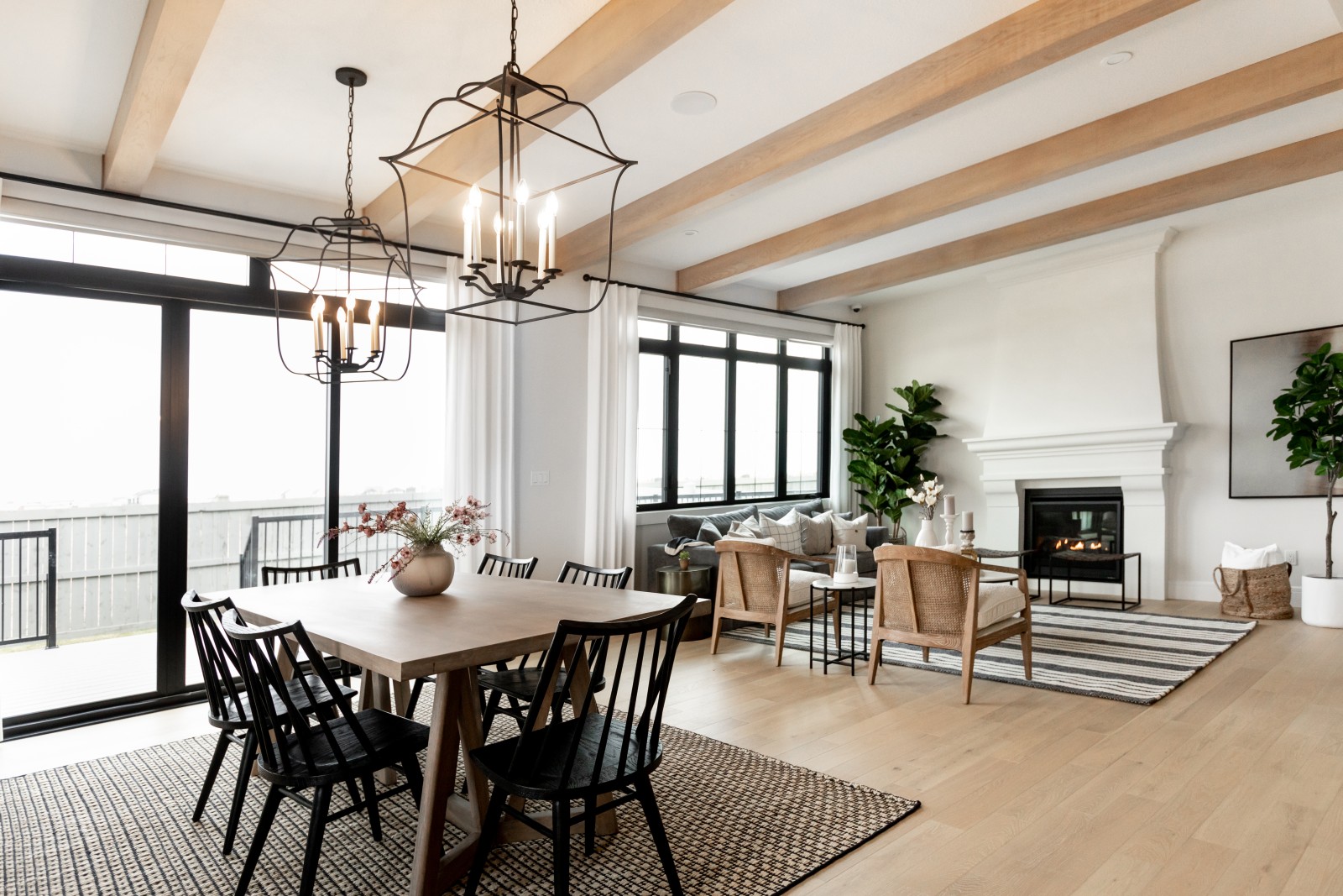 A wider shot of the main floor of the Kalliope Griesbach showhome featuring warm wood beams running across the space. Two large black chandeliers hang above the wood dining table with accenting black chairs. The inviting living room with ten foot tall custom plaster fireplace can be seen in the back of the photo