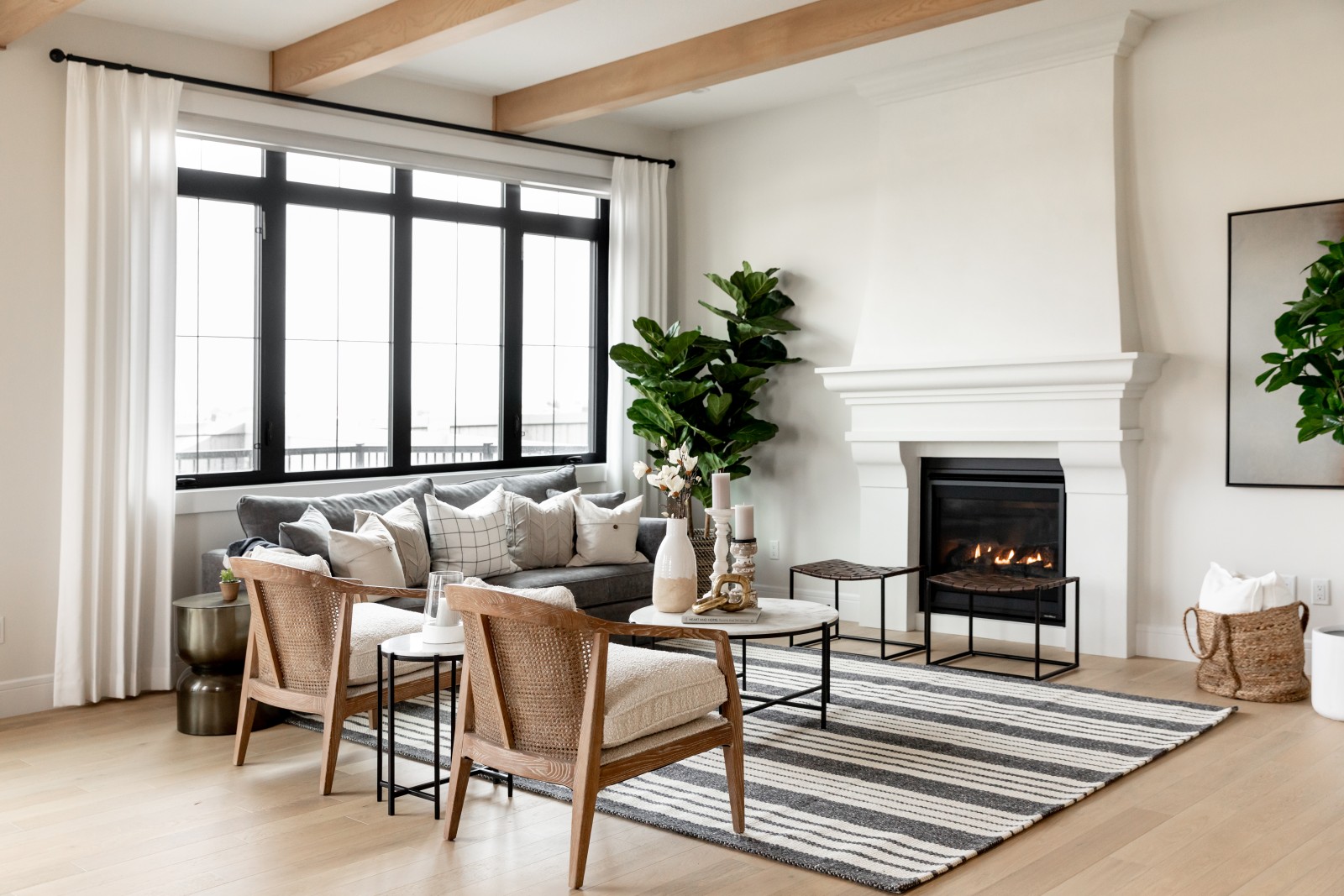 A ten foot tall, custom plaster fireplace, is the focal point of this photo. A cozy couch sits below the black framed windows found at the back of the home and two warm wood chairs sit across from the fireplace