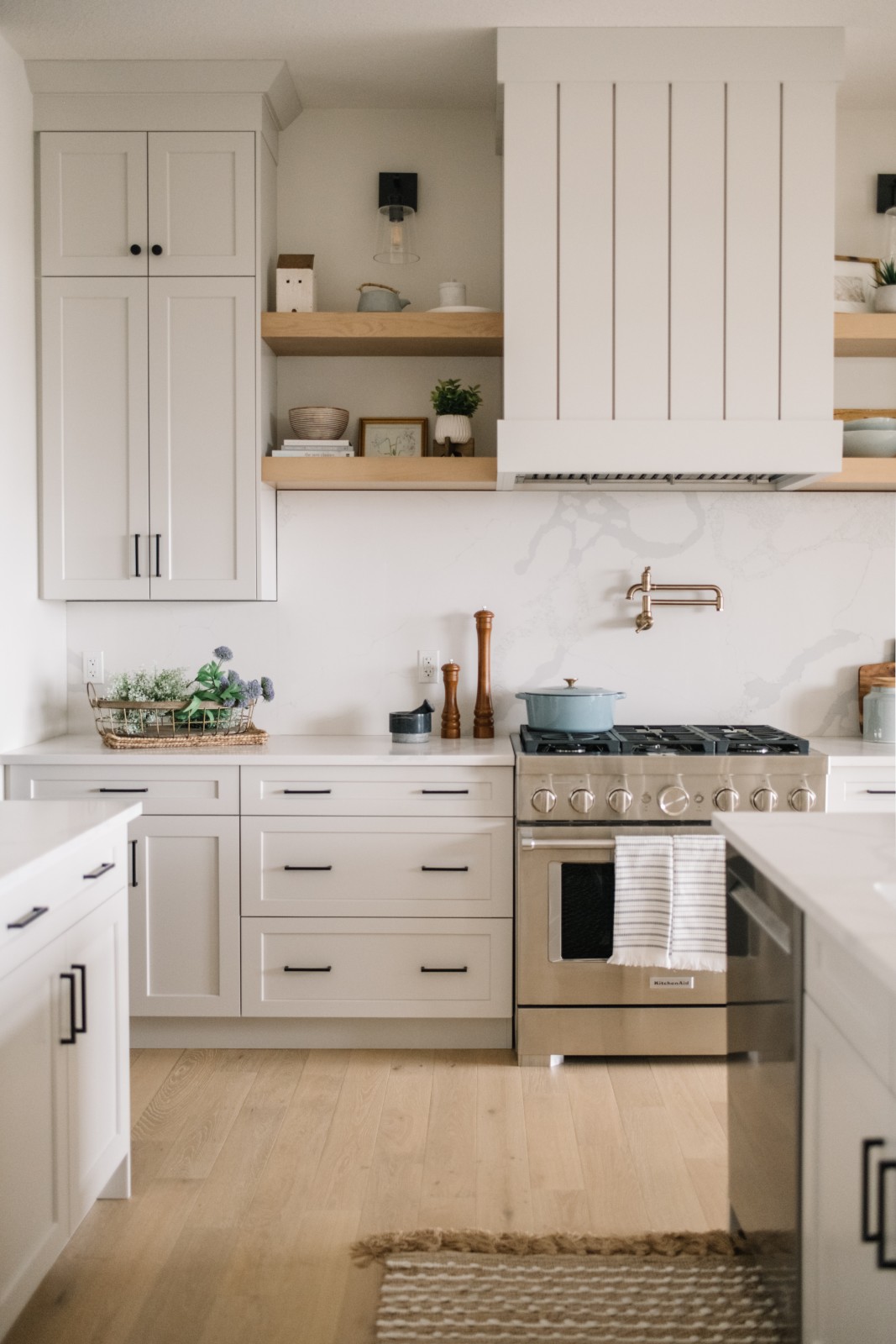 Range wall on of the Kalliope showhome in Griesbach featuring a custom vertical shiplap wood hood fan, flanked by warm wood floating shelves and stacked griege cabinets to the side. The backsplash is a solid piece of quartz countertop with grey veining carried up from the countertop.