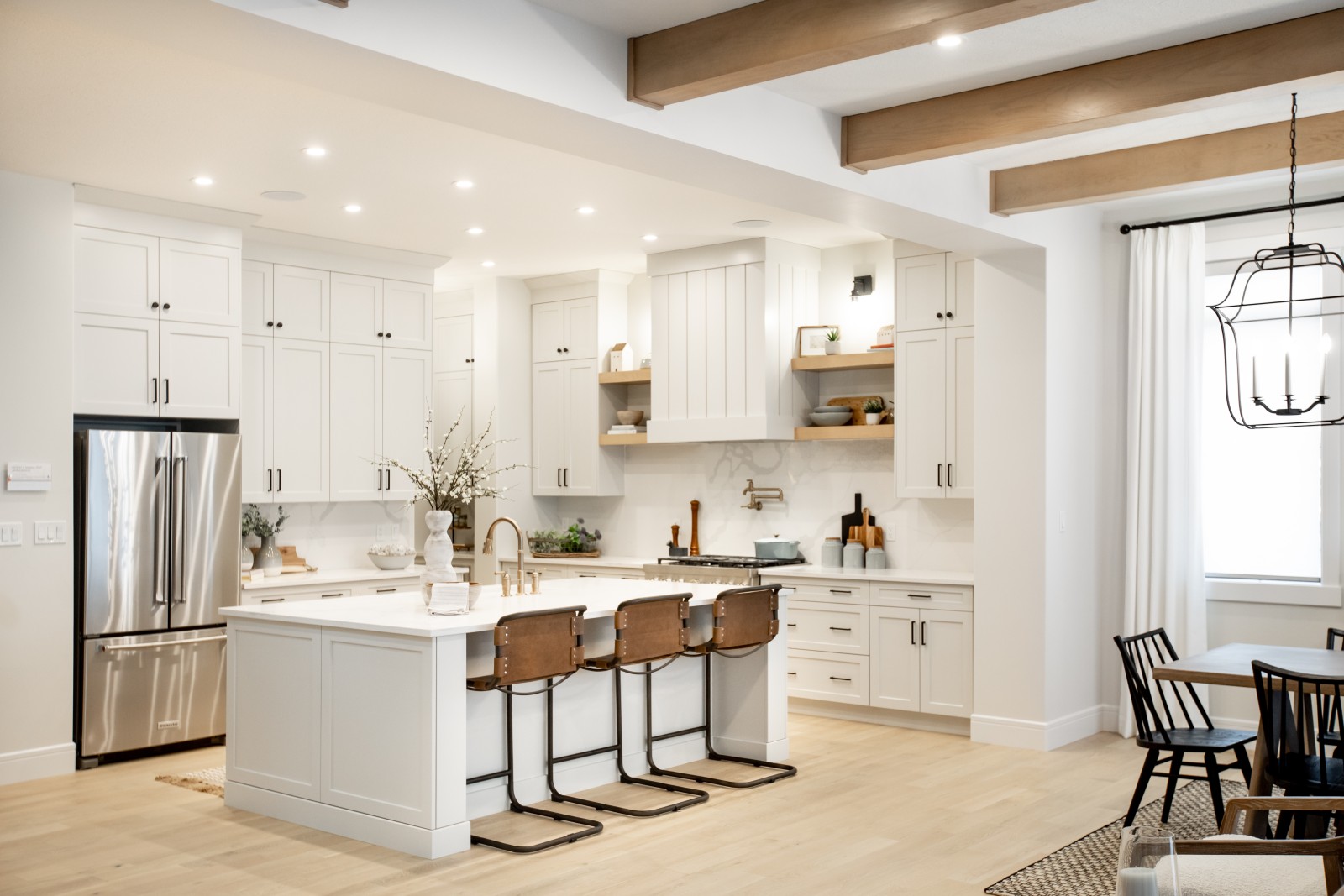 Wide shot of the bright, light and warm kitchen of the Kalliope showhome with light beige cabinets, white and grey veined quartz countertops and backsplash, accents of natural wood in the floating shelves flanking the custom vertical shiplap hood fan. The warm wood beams are also visible on the ceiling of the dining room next to the kithcen