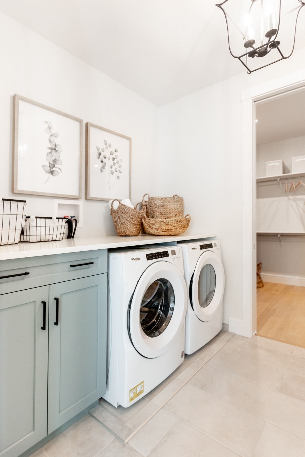 Light blue cabinetry in the Kalliope Griesbach showhome laundry room with white countertop above front load appliances. Natural light art with wood frames hang on the wall and compliment the wicker baskets on the countertop