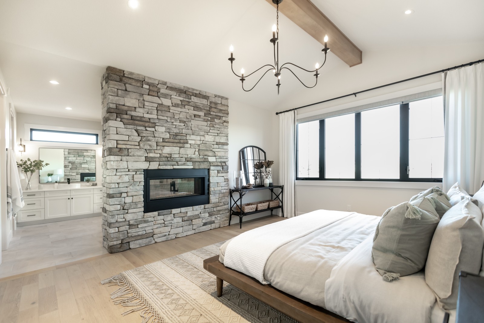 A view of the ensuite from the master bedroom of the Kalliope showhome. The bedroom has a warm wood beam running across the room, with a large iron chandelier hanging from it. The dual sided fireplace, with stone from floor to ceiling, separates the bedroom from the ensuite.