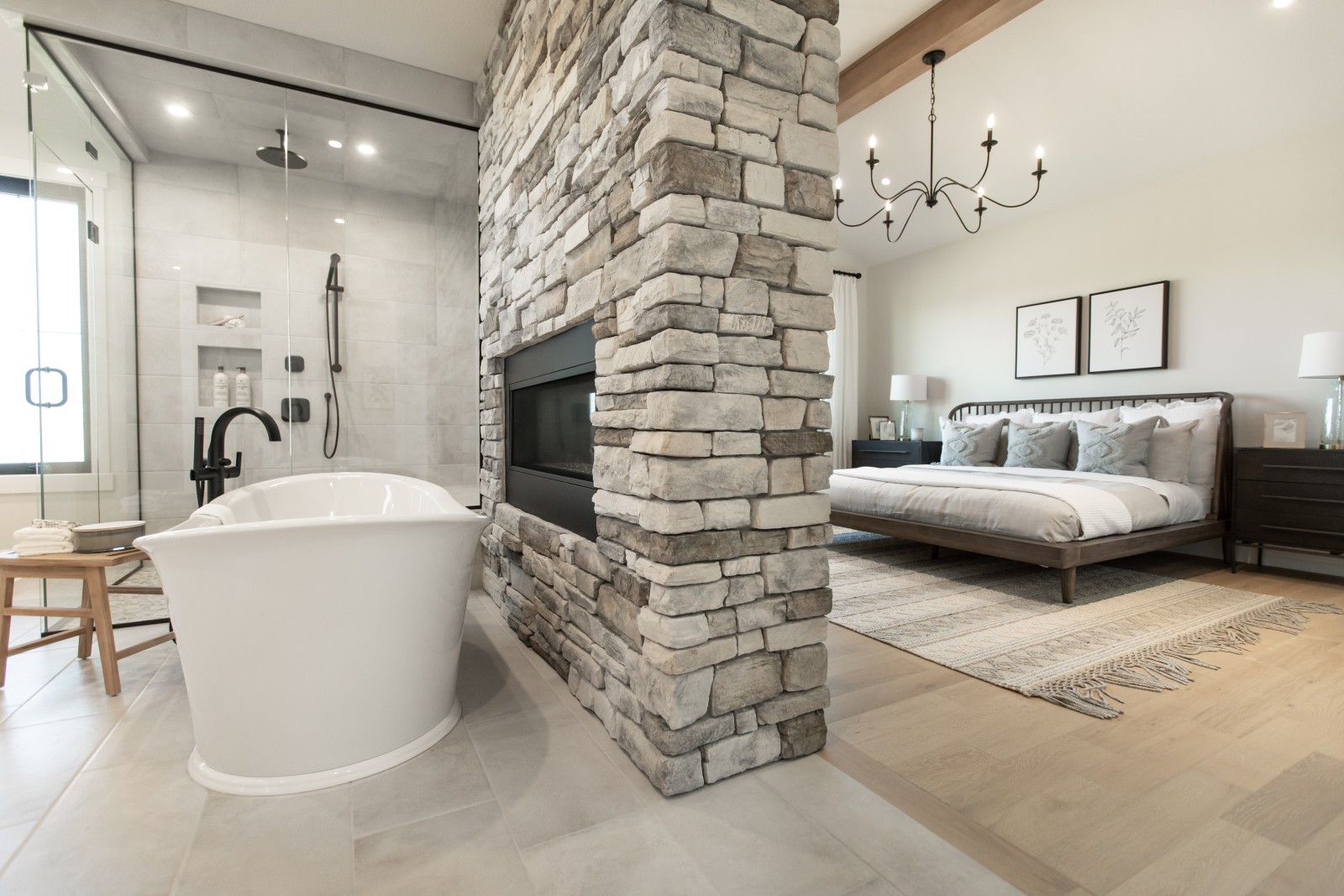 A view of both the ensuite and the master bedroom with the dual sided fireplace, which has stone from the floor to the ceiling, sitting at the center of the photo. The cozy, warm wood platform bed with white bedding can be seen behind the fireplace. The freestanding tub and fully tiled shower can be seen in front of the fireplace