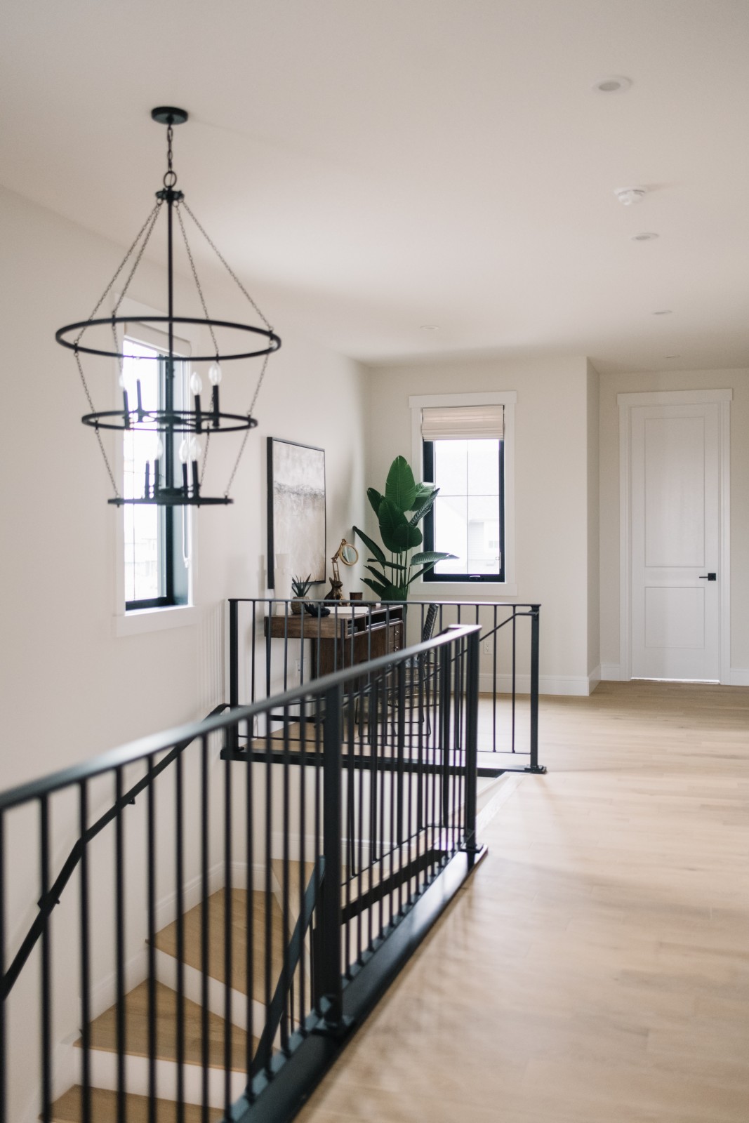 A large black, tiered chandelier hangs above the stairwell leading up to the second floor of the Kalliope showhome complimenting the iron railing. Hardwood floors of the second floor hallway lead to the office nook located at the top of the stairs