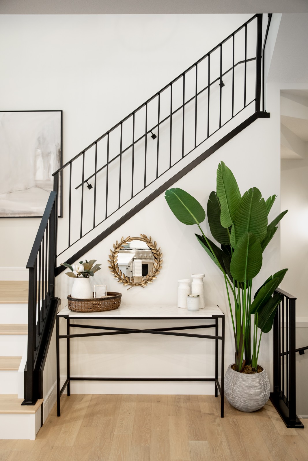 Black iron railing leading from the main floor to the second pop off of the warm white walls. In the nook below the railing a side table is accessorized with a small gold mirror on the wall, small white and natural vases and a large leafed floor plant