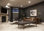 The main living room in the basement with dark, moody wall, a fireplace in the corner with a wood mantle and stone below the mantle, an inviting cognac leather couch on the far wall and light art hanging on the wall