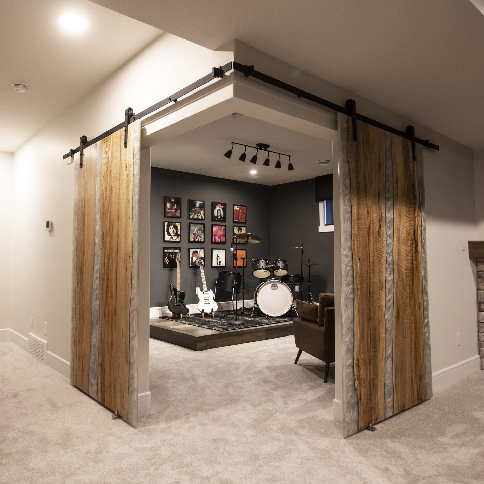 Two wood and epoxy barn doors opened to reveal a music room with dark walls, a stage in the corner, a drum set and guitar on the stage, a musical art collage hanging on the wall behind the instrument and two chairs across from the stage for viewing