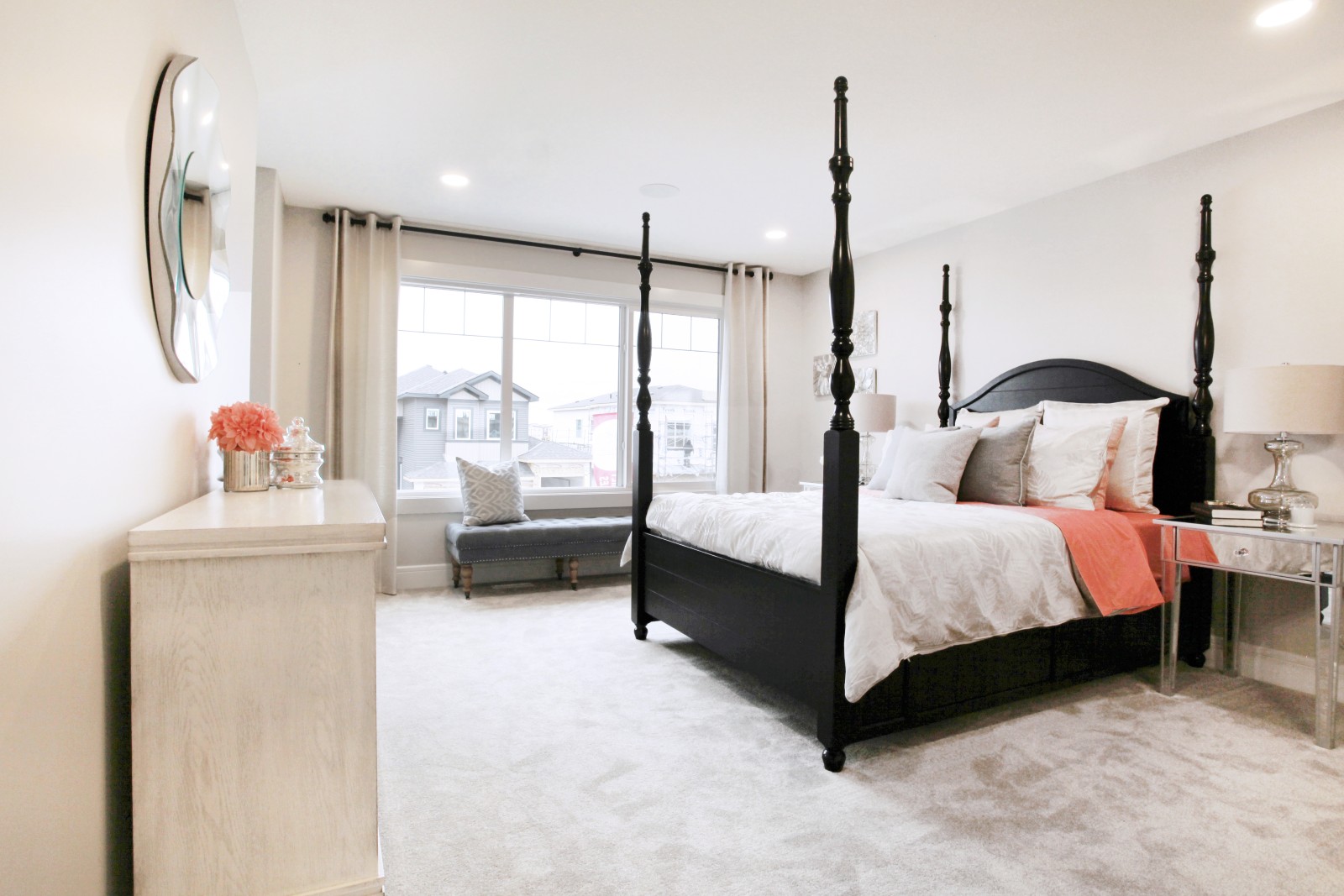 Master bedroom of the Julian showhome with large windows at the front of the room, a dark four-poster bed with light bedding and pops of coral accents