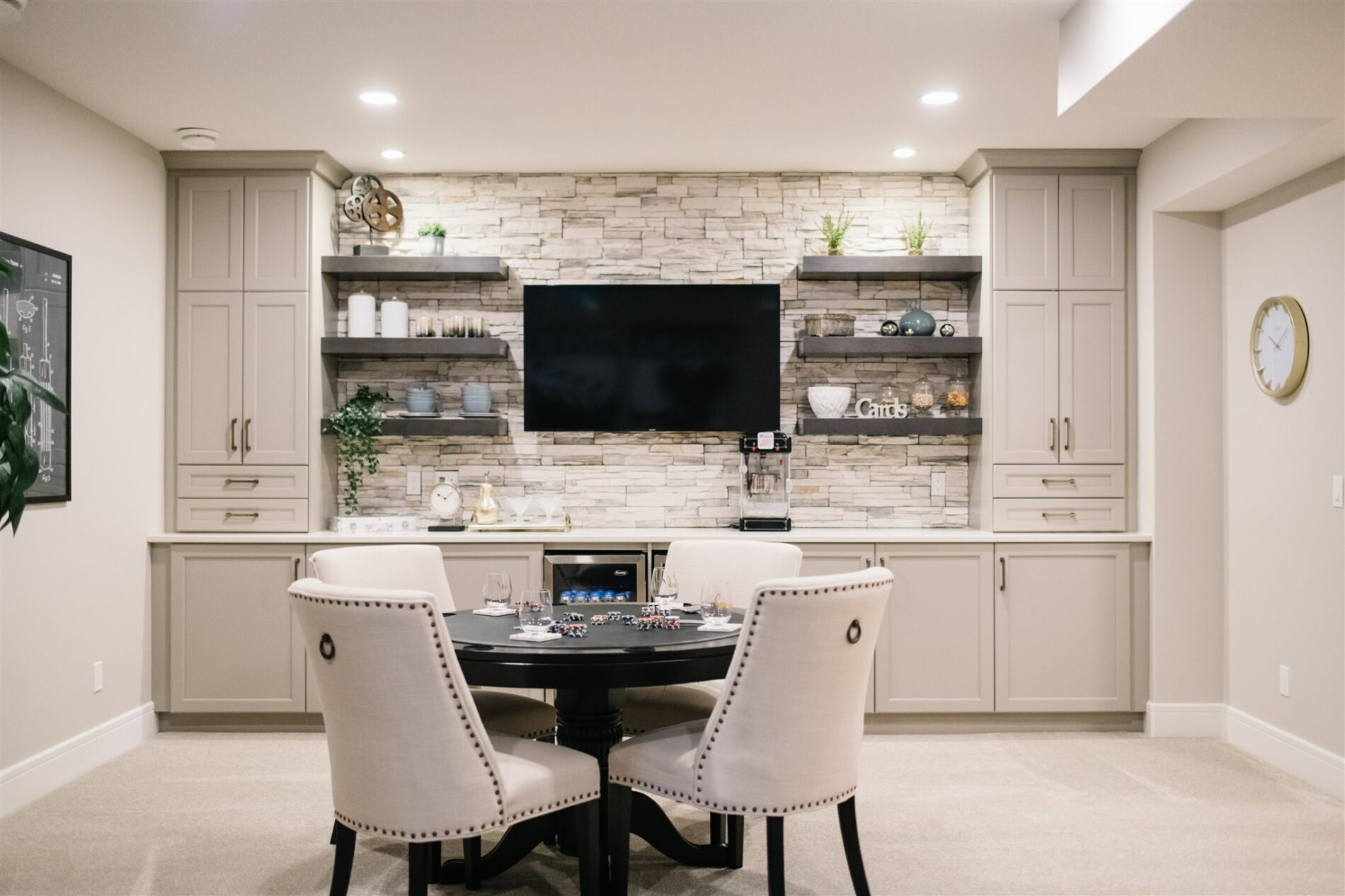 Games table set up in front of dry bar in basement of the Julian showhome with light beige cabinetry, stone feature wall and floating shelves infront