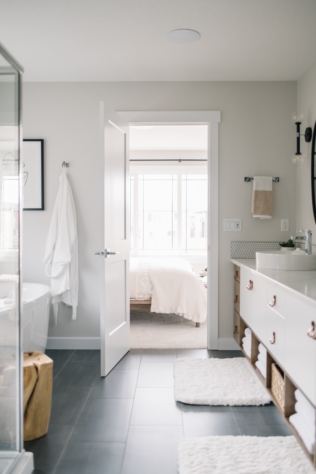 A view of the ensuite looking in to the master bedroom of the Nysa showhome in Cy Becker. The dark floor tile is brightened by all white wall, a long white and warm wood dual vanity with white countertops and round vessel sinks, and the white freestanding tub in front of the window