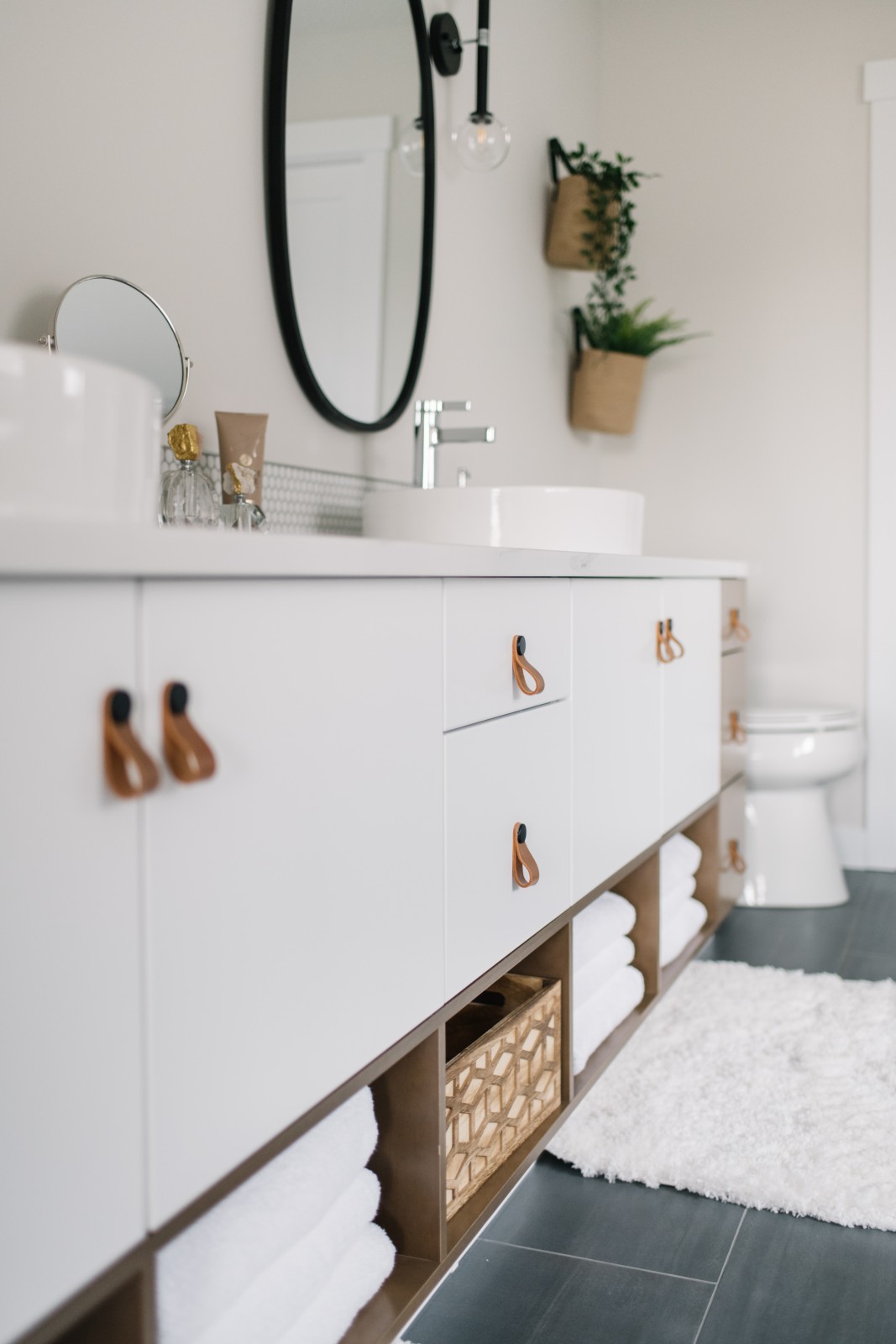 A long sleek white vanity with cognac leather pulls and coordinating wood shelves. Large black framed, oval mirrors can be seen hanging above the vessel sinks on the white countertops