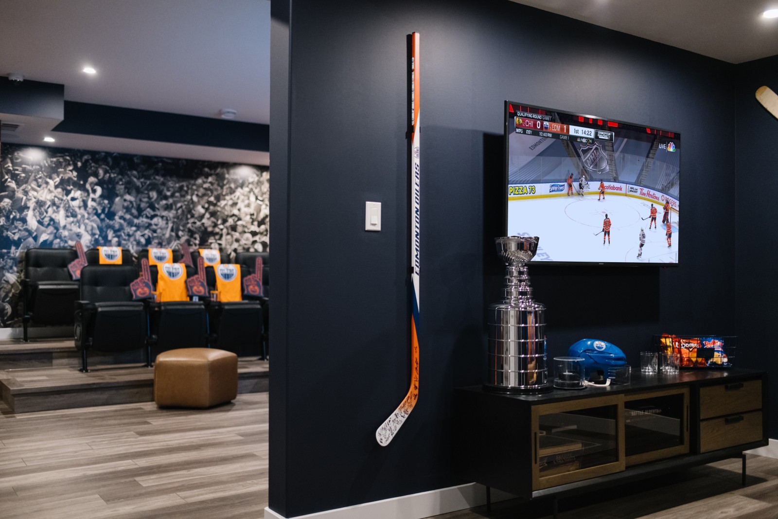 Oilers Fan Cave in the basement of the Nysa showhome in Cy Becker. A large tv and hockey stick are mounted to the dark blue wall. Stadium seats in front of a mural of a crowd in the arena can be seen behind