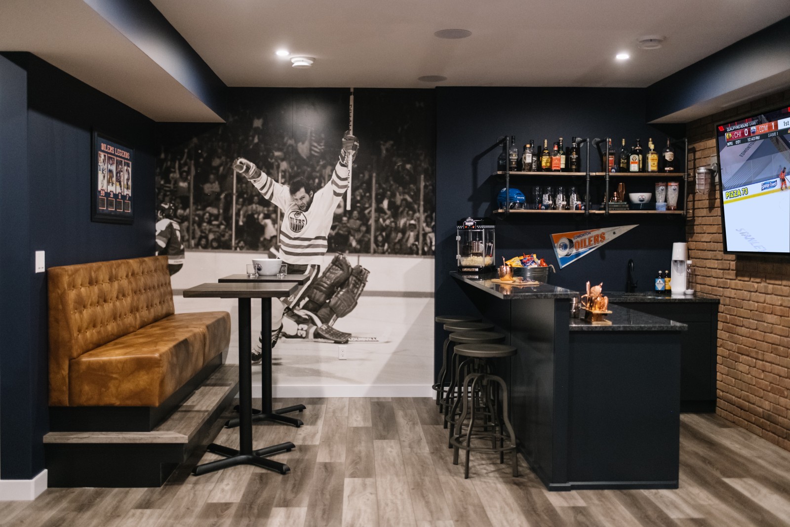Oilers Fan Cave in the Nysa showhome with a floor to ceiling, black and white mural of a hockey player celebrating. A cognac leather banquet and bar tables are to the left of the mural and a bar with dark blue cabinetry and floating shelves are to the right of the mural.