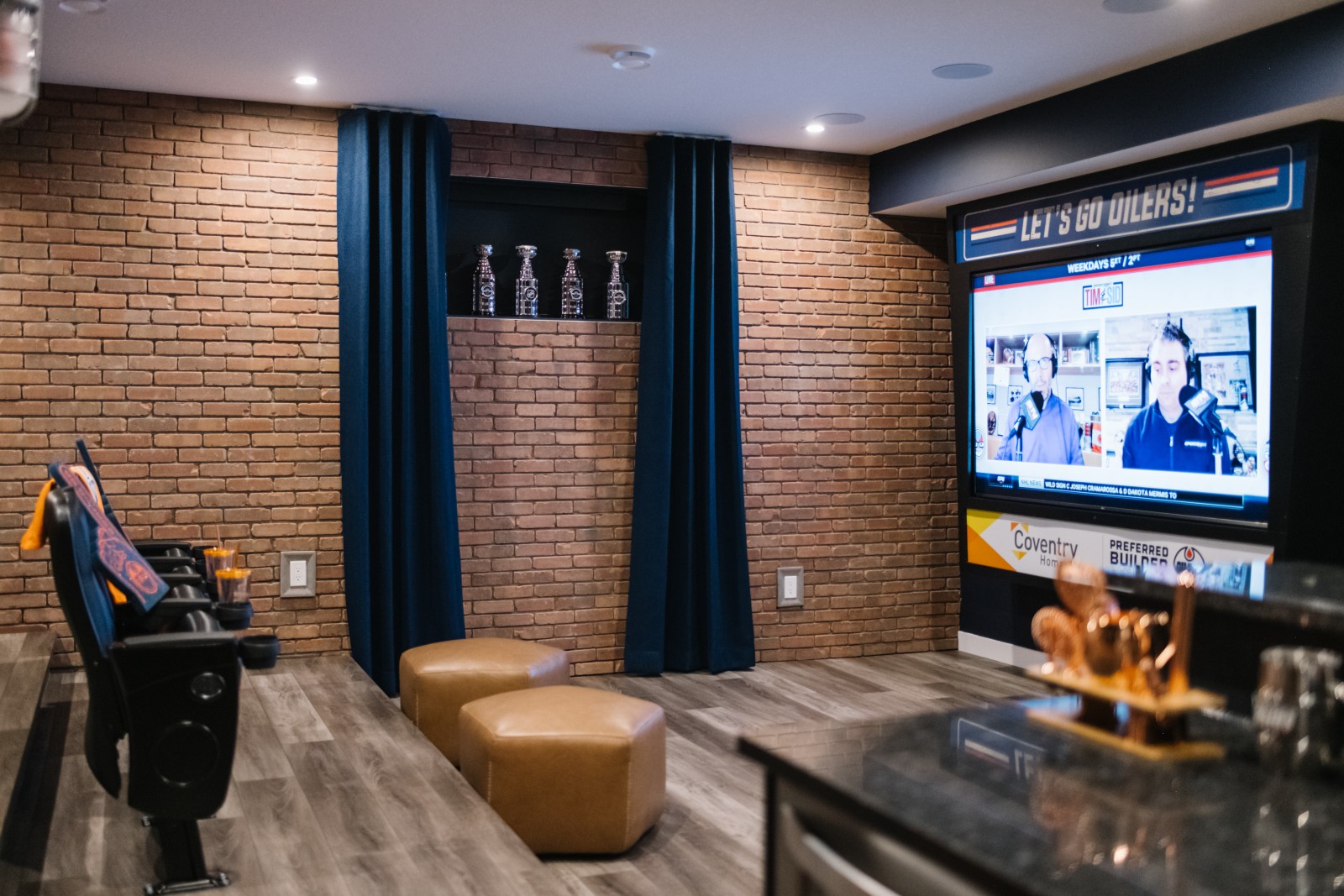 Oilers Fan Cave in the Cy Becker showhome. A brick wall is the focal point of the photos with a large wall mounted jumbotron to the right of the wall and stadium seats to the left