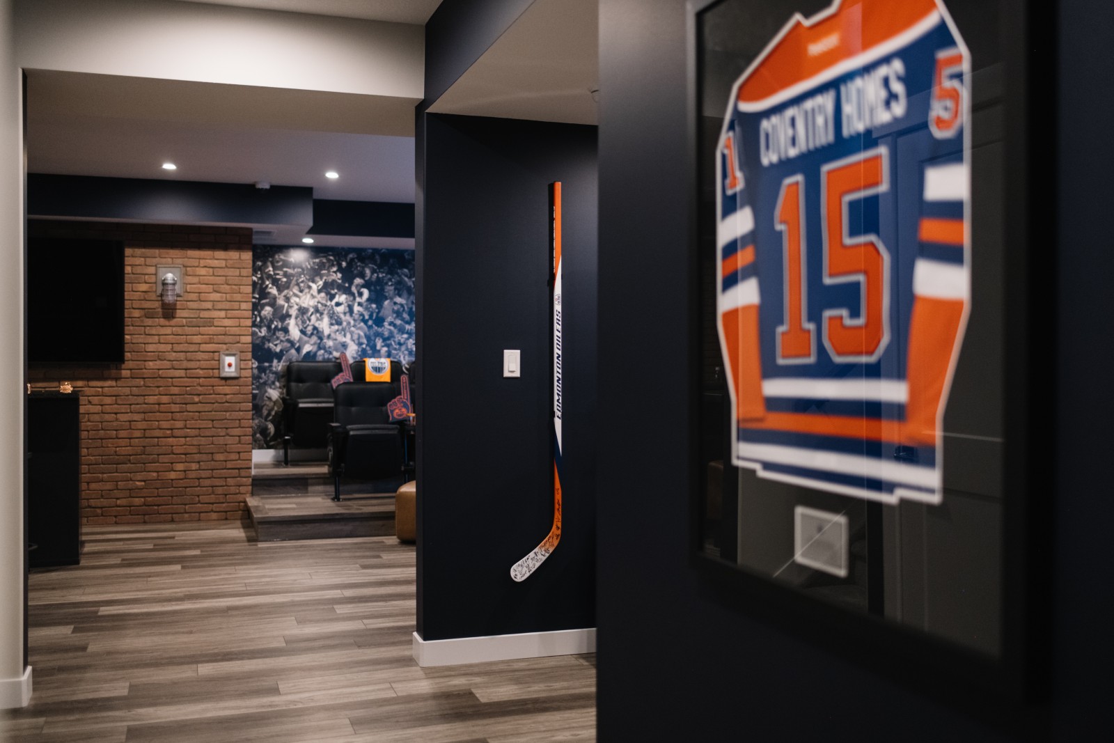 Oilers Fan Cave in the basement of the Nysa showhome. A framed Oilers Jersey is hanging on a dark blue wall. Behind you can see a tv hanging on a brick wall and stadium seats in front of a mural of a crowd of people