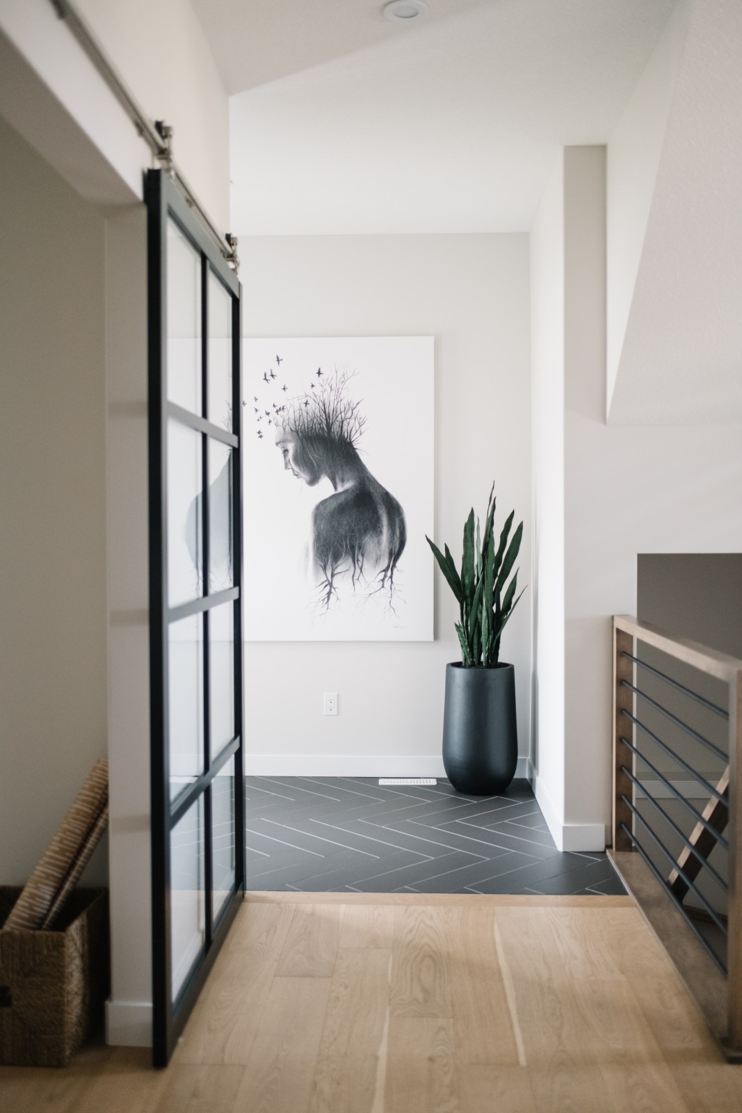 The front foyer of the Nysa showhome with black herringbone floor tile, white walls, modern black and white art of a woman. A black and glass barn door can be seen leading to a room to the left