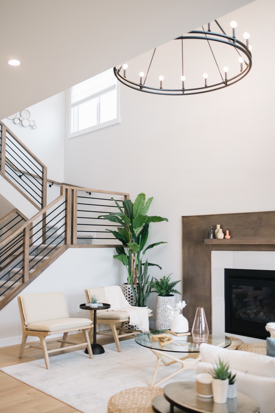 Looking at the open to above great room of the Nysa showhome, a large round, black iron chandelier can be seen in the top corner of the photo. The fireplace with warm wood surround is to the right of the stained wood railing with black horizontal spindles leading to the second floor.