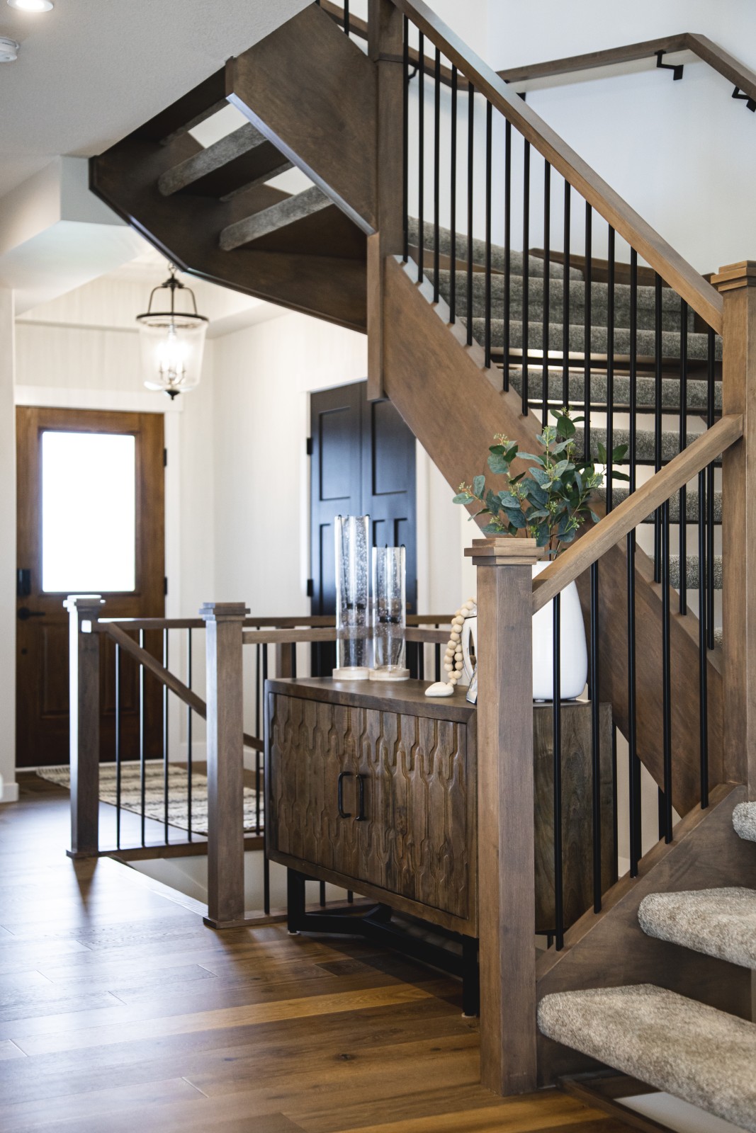Stained wood staircase with black spindles and open treads. The front door behind the staircase and sideboard in the nook of the staircase are in complementing warm wood tones