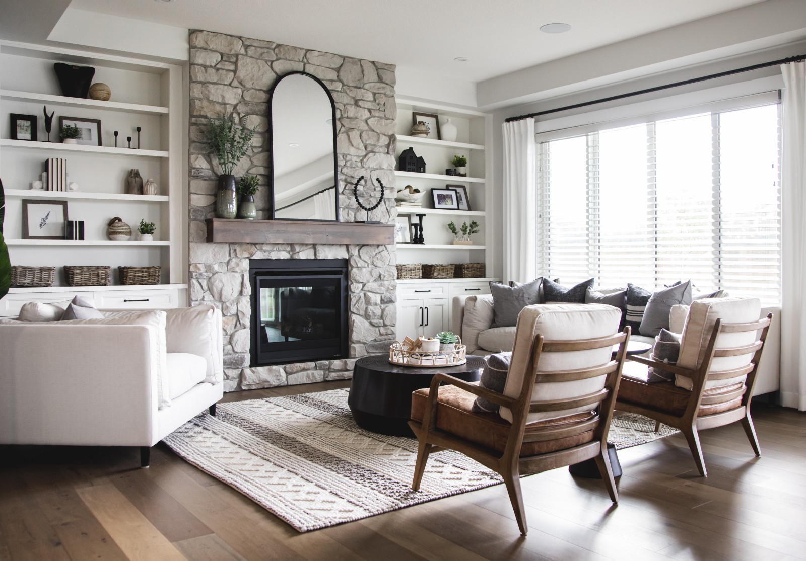 A fireplace clad in light grey stone from floor to ceiling with a warm wood mantle is the focal point of the photo. A cozy couch is placed to the side of the fireplace in front of the large window in the back of the home. Wood accents are carried in to the chairs seated opposite of the fireplace.