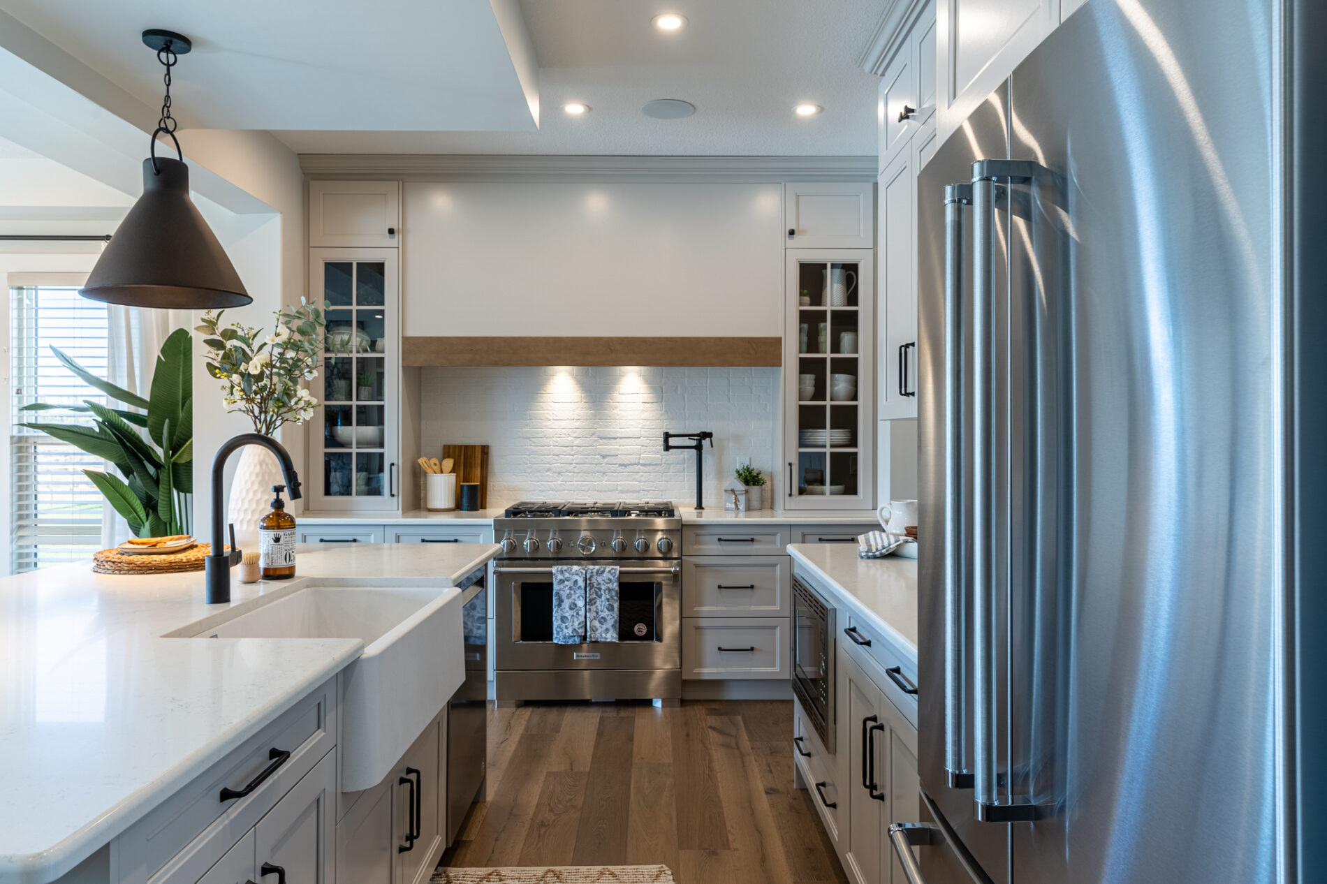 An extended custom wood hood fan with warm wood trim along the bottom is the focal point of the range wall in the Odessa Showhome. Glass cabinets flank the range and are complimented with white, textured backsplash tile and white countertops.