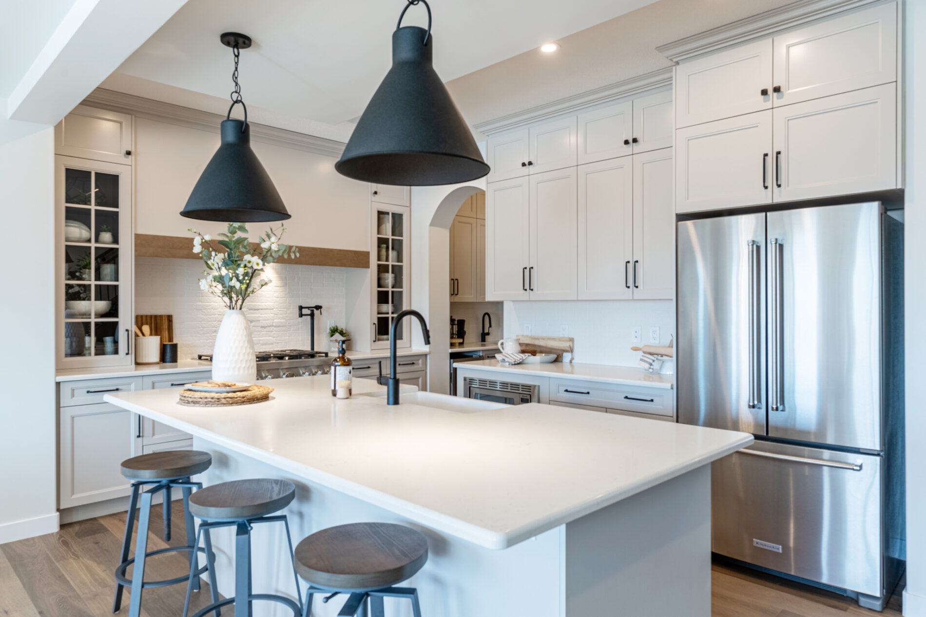 Two large black light fixtures are centered over the flush eating bar in the island of the Odessa Showhome. Black accents in the cabinets handles and stools compliment the light and airy kitchen.