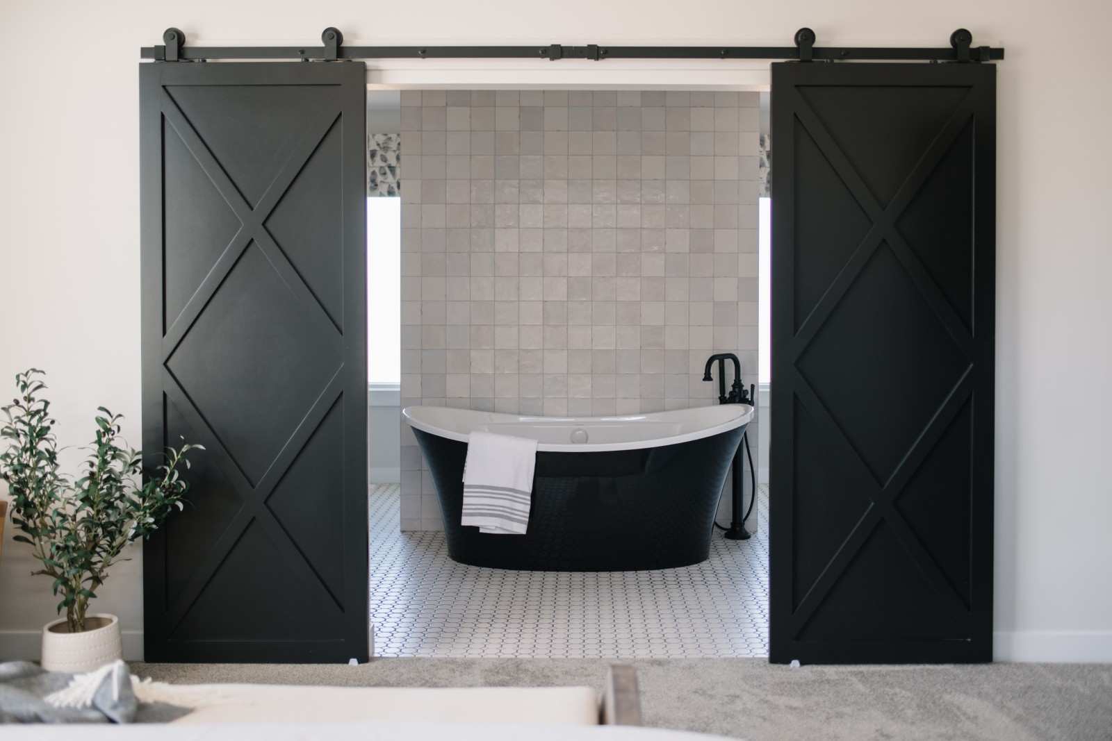 Black barn doors open to reveal a black free standing tub in front of a wall with floor to ceiling, light coloured, textured tile in the Odessa showhome in Riverside