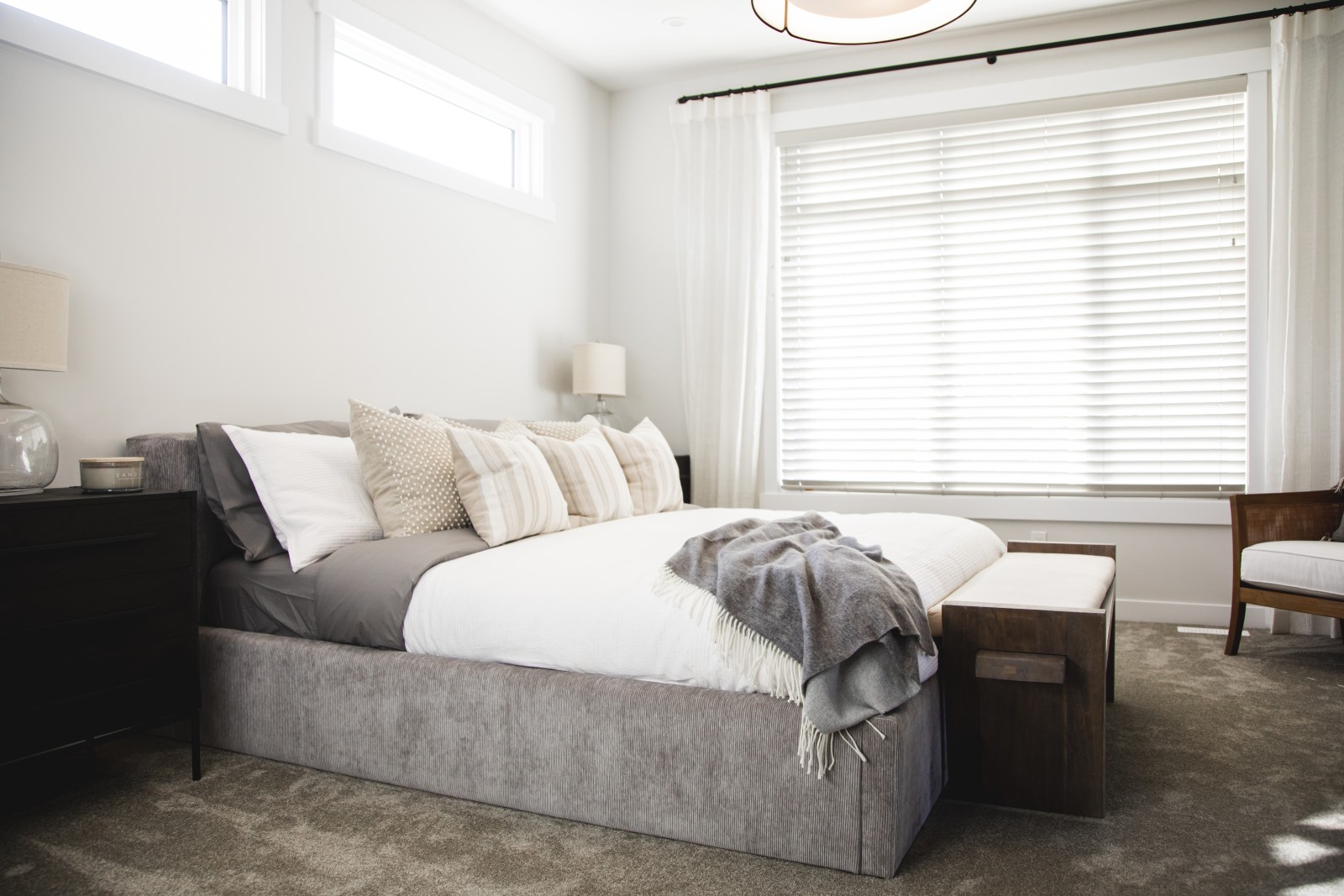 Natural light floods the master bedroom of the Odessa showhome through a wall of windows at the back of the room as well as two piano windows above the bed. An upholstered, grey platform bed is centered on the wall