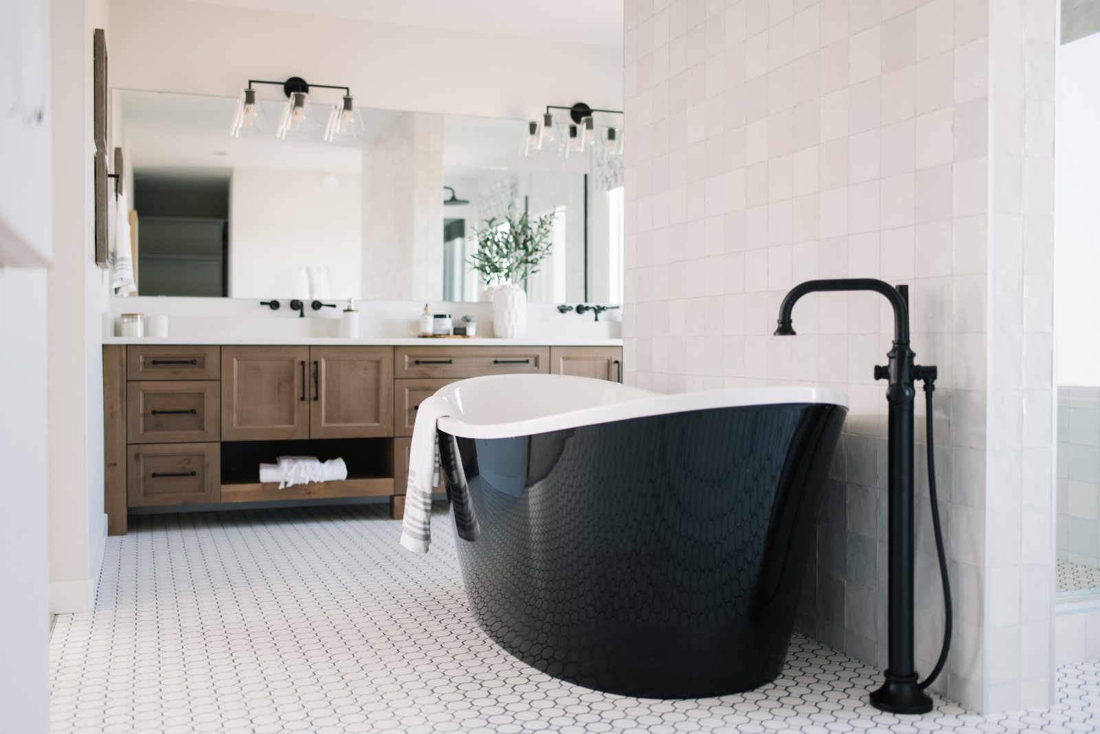 A warm and inviting ensuite with black soaker tub in the center of the room infront of a wall of simple textured tile. A warm wood can be seen at the back of the room with open shelving for storage, white countertops, vessel sinks and wall mounted, black faucets.