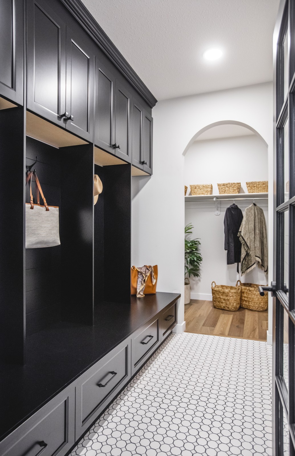 Back entry to the Odessa showhome with beautiful black built in bench and cubbies with matching cabinets above for storage. Black and white patterned tile floor leads to a walk-in closet with hardwood floors and hanging shelf, separated from the back entry by an arch. A black framed glass door can be seen on the right of the photo.