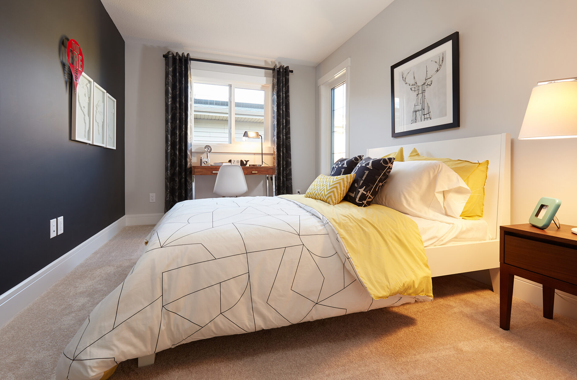 Simplistic secondary bedroom with bold black feature wall, white and black patterned bedding, black curtains, with a desk below the window and yellow accents in bedding and pillows