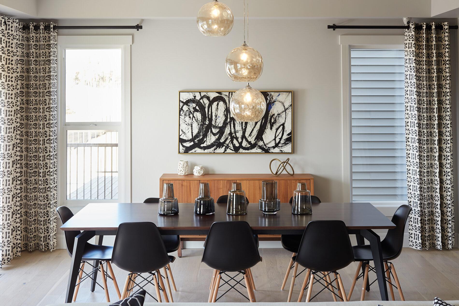 Dining room in Kalliope showhome with long dark wood table, eight black Eiffel chairs, three globe light fixtures hung at different heights above the table, a large warm wood side table against the wall between two large windows and a large piece of black and white art hanging on the wall