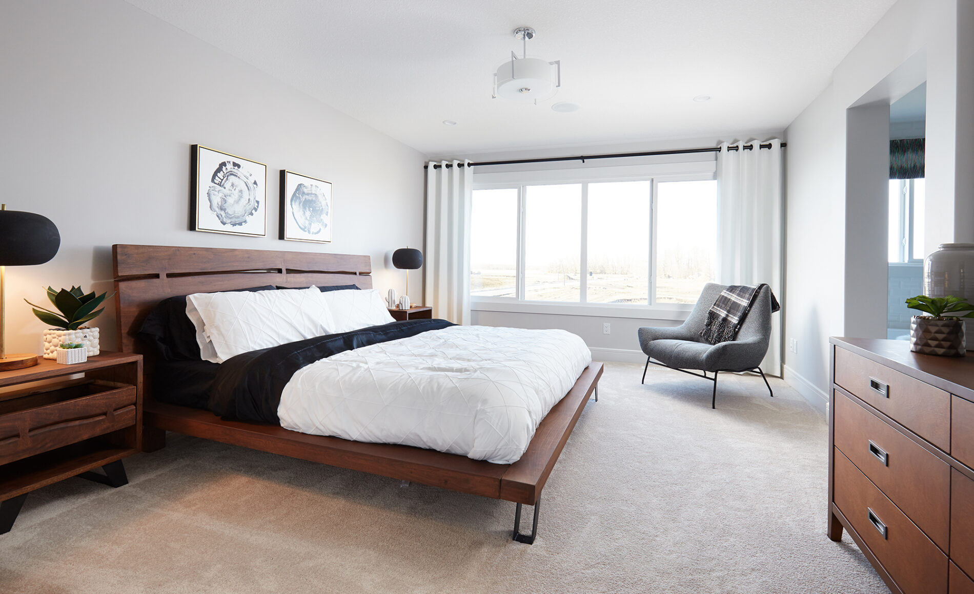 Bright master bedroom with warm wood platform bed, white and black bedding, art above the bed and a chair in the corner in front of large windows at the back of the room