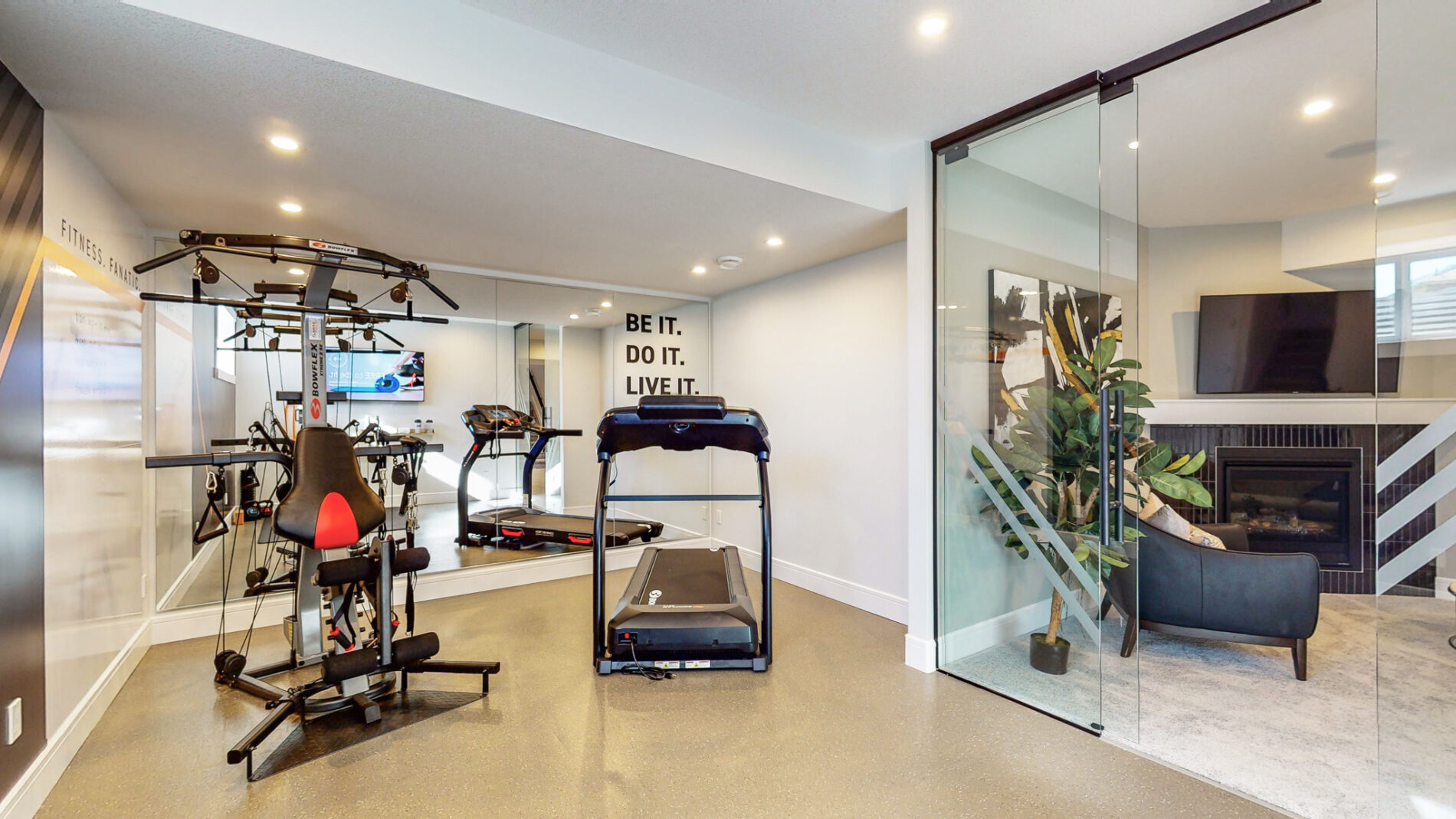 Home gym in the Kail showhome with glass wall separating the gym from the family room, light rubber flooring, floor to ceiling mirror on one wall and gym equipment centered in the room