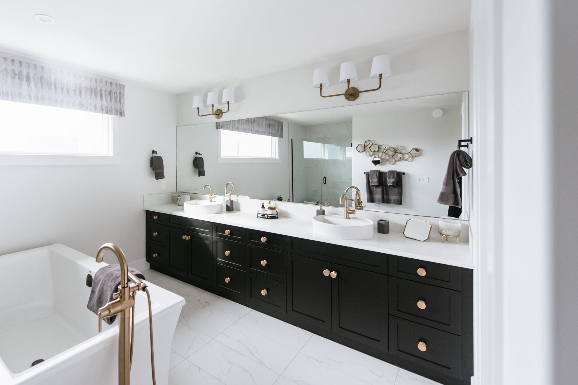 Master ensuite in the Kail showhome featuring luxurious black dual vanity with large gold handles, vessel sinks with gold faucets, a free standing tub in the middle of the room with a gold tub filler and the fully tiled shower reflecting in the mirror