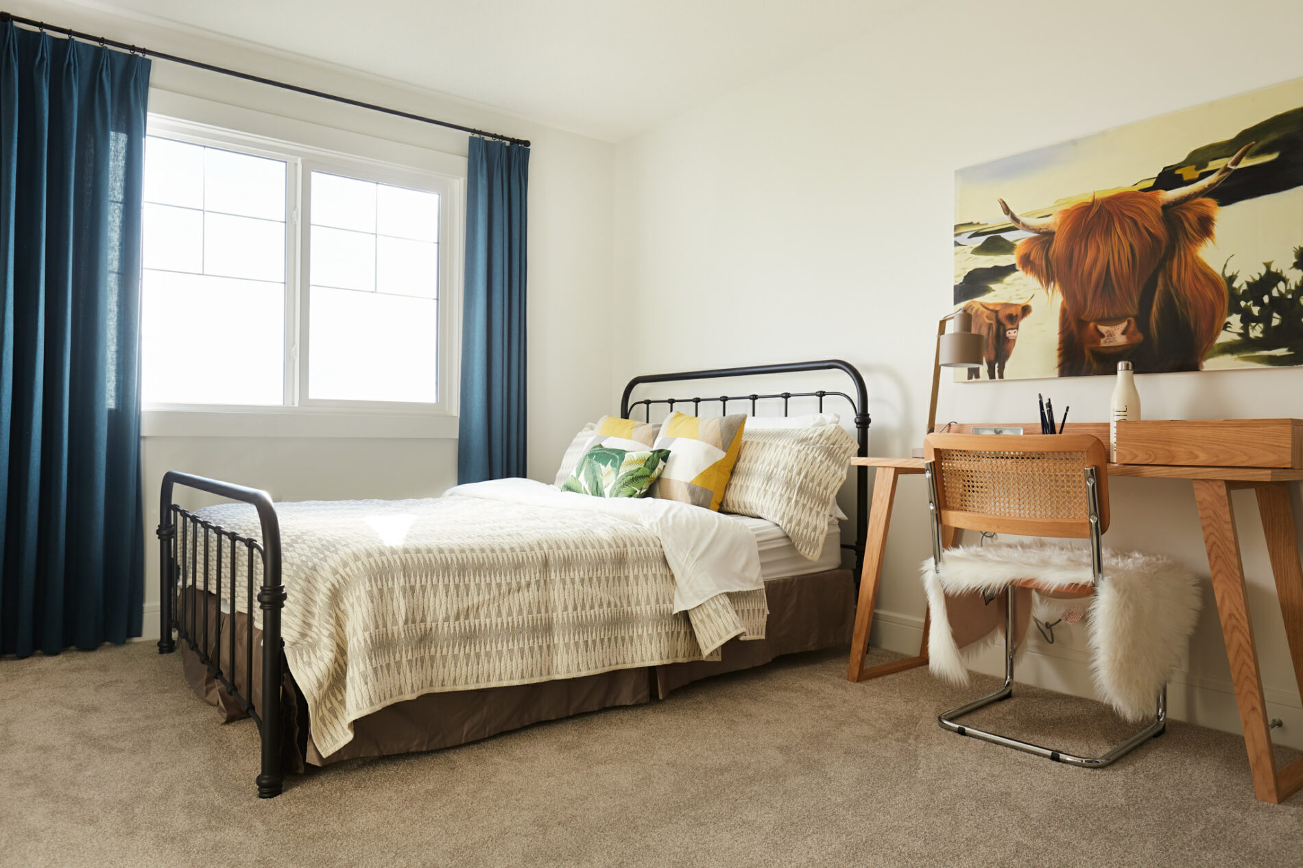A gender neutral secondary bedroom with large window, wrought iron bed with rustic bedding, wood desk and chair and large highland cow art