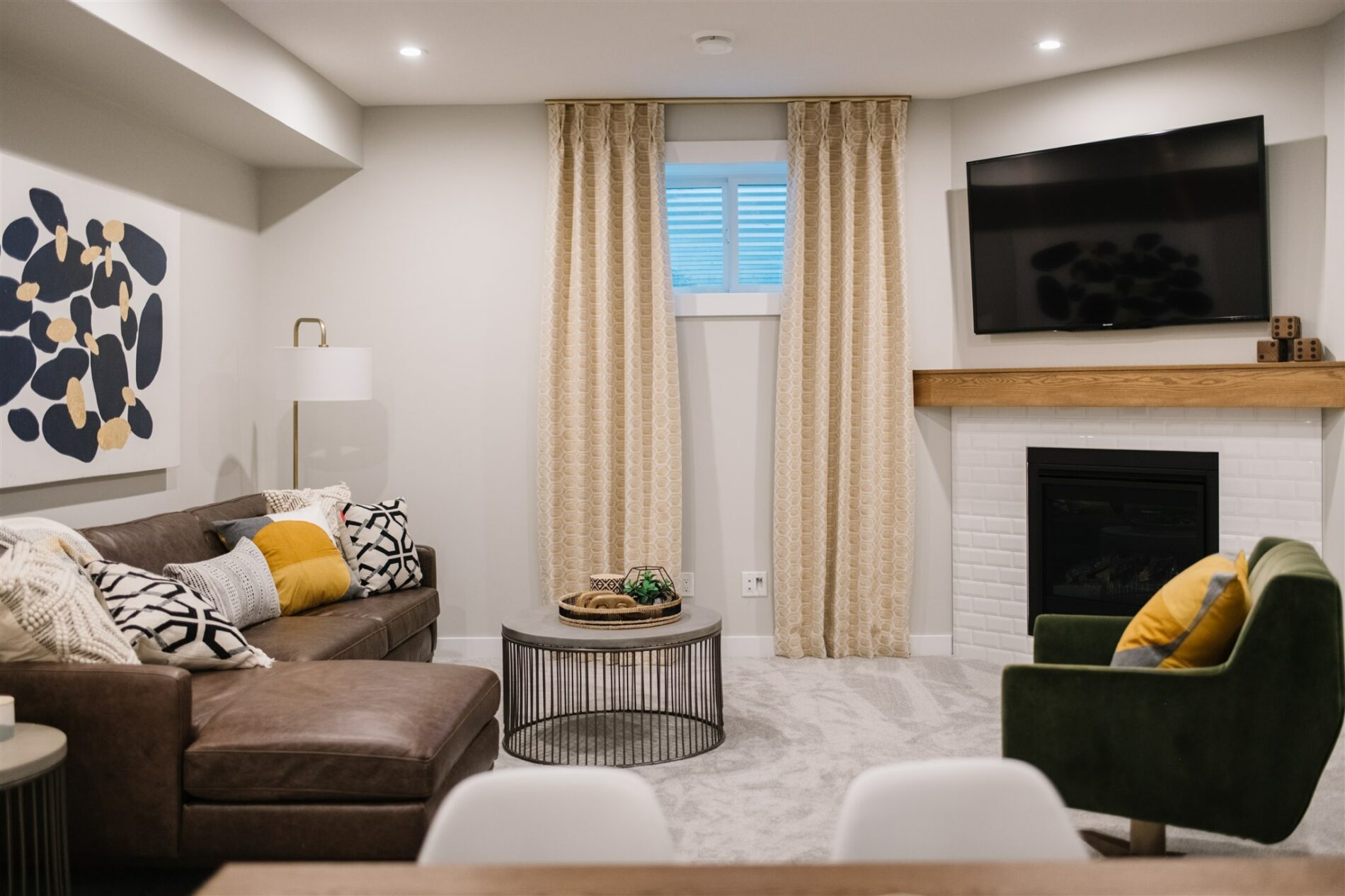 A cozy basement with corner fireplace with stained wood mantle, tv mounted above, warm gold curtains and rich, dark leather couch