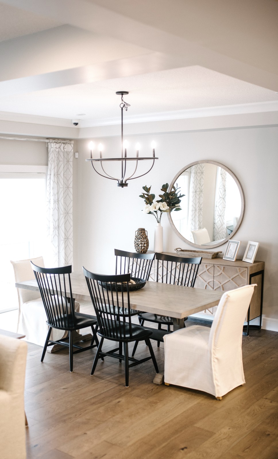 A classic dining room with wood table and black high back wood chairs with white covered chairs at the heads of the table, classic sideboard and oversized round mirror hung above on the wall