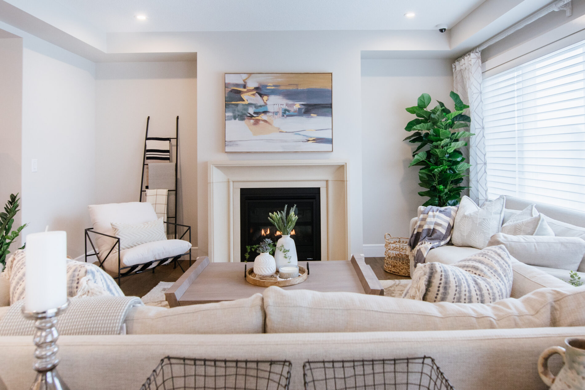 Warm and inviting, light and bright Great Room with fireplace with stone mantle as the central focal point of the room. Light and white furniture is placed around the fireplace accessorized by a tree in the corner by the window, a blanket ladder to the left of the fireplace and an large painting above the fireplace