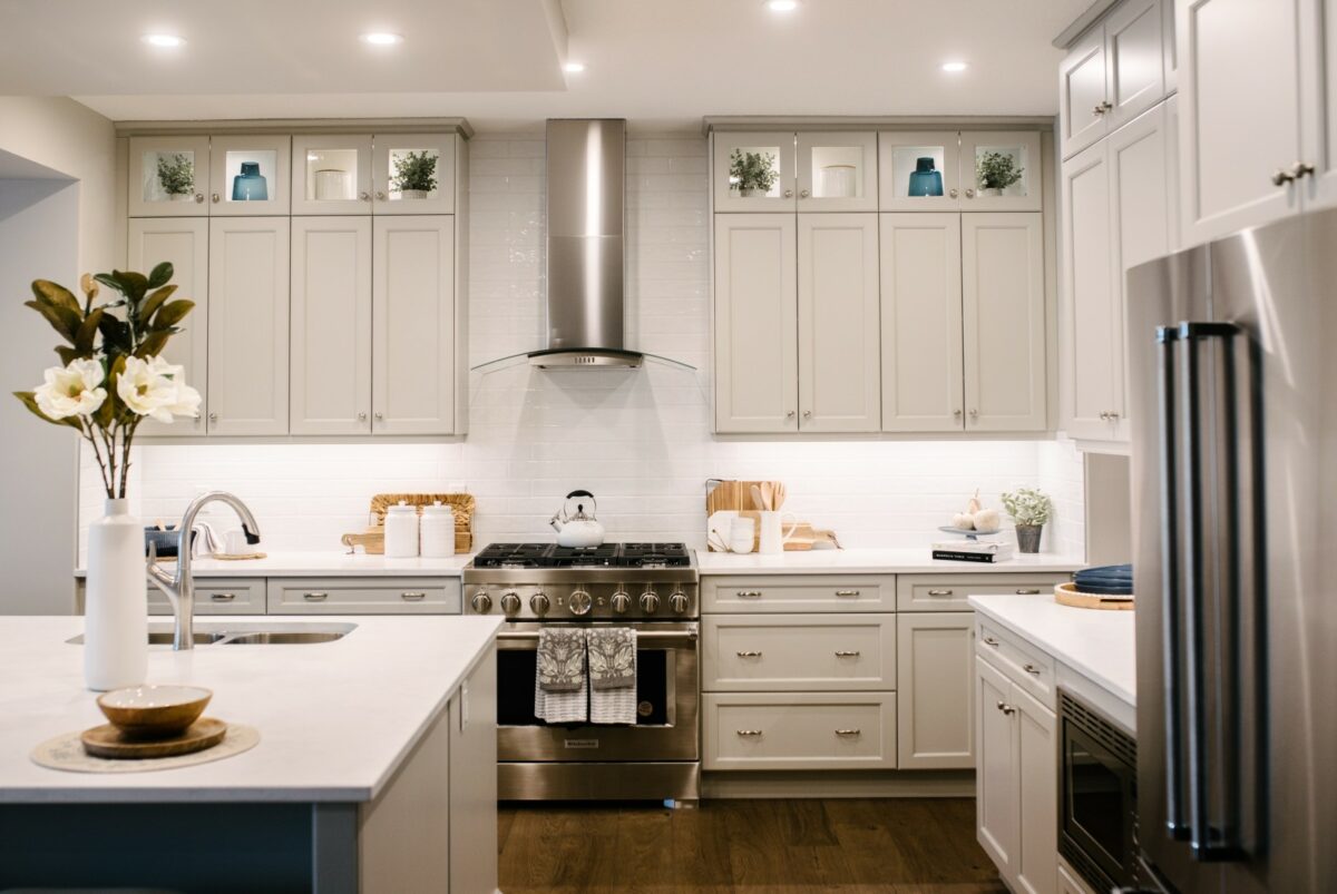 The range wall of a kitchen with light grey/beige stacked cabinets, white backsplash and countertops, stainless steel appliances, accessorized with warm wood