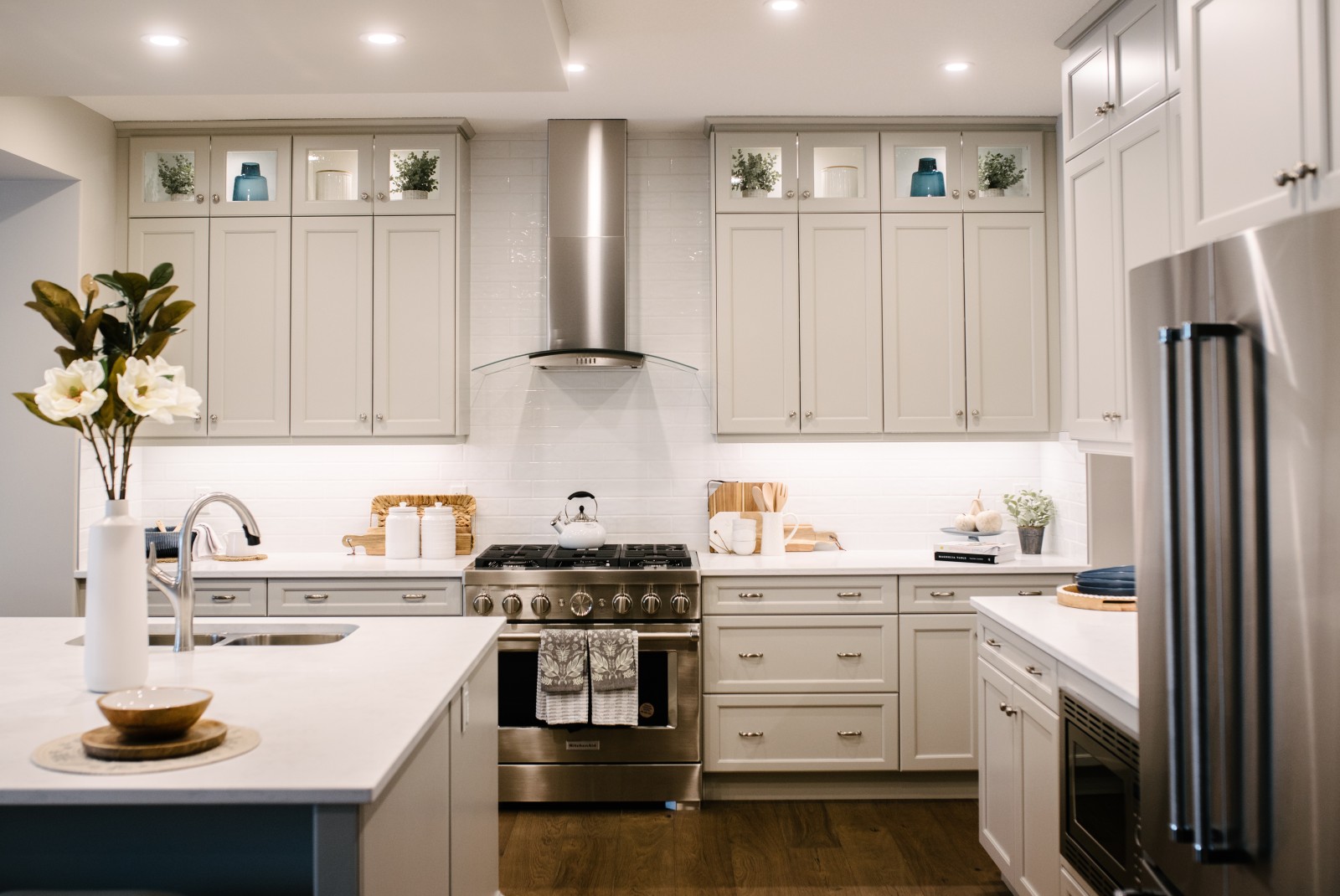 The range wall of a kitchen with light grey/beige stacked cabinets, white backsplash and countertops, stainless steel appliances, accessorized with warm wood