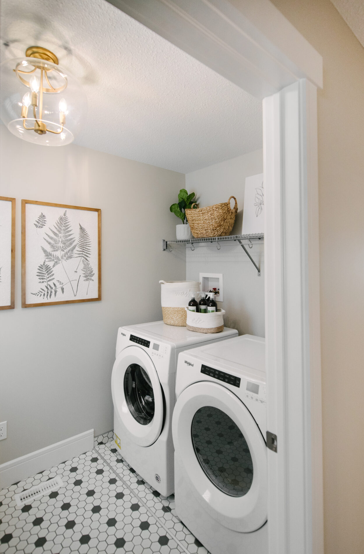 A bright laundry room with black and white patterned floor tile, front load appliances, a shelf above and some accessories in natural finishes such as wood and wicker