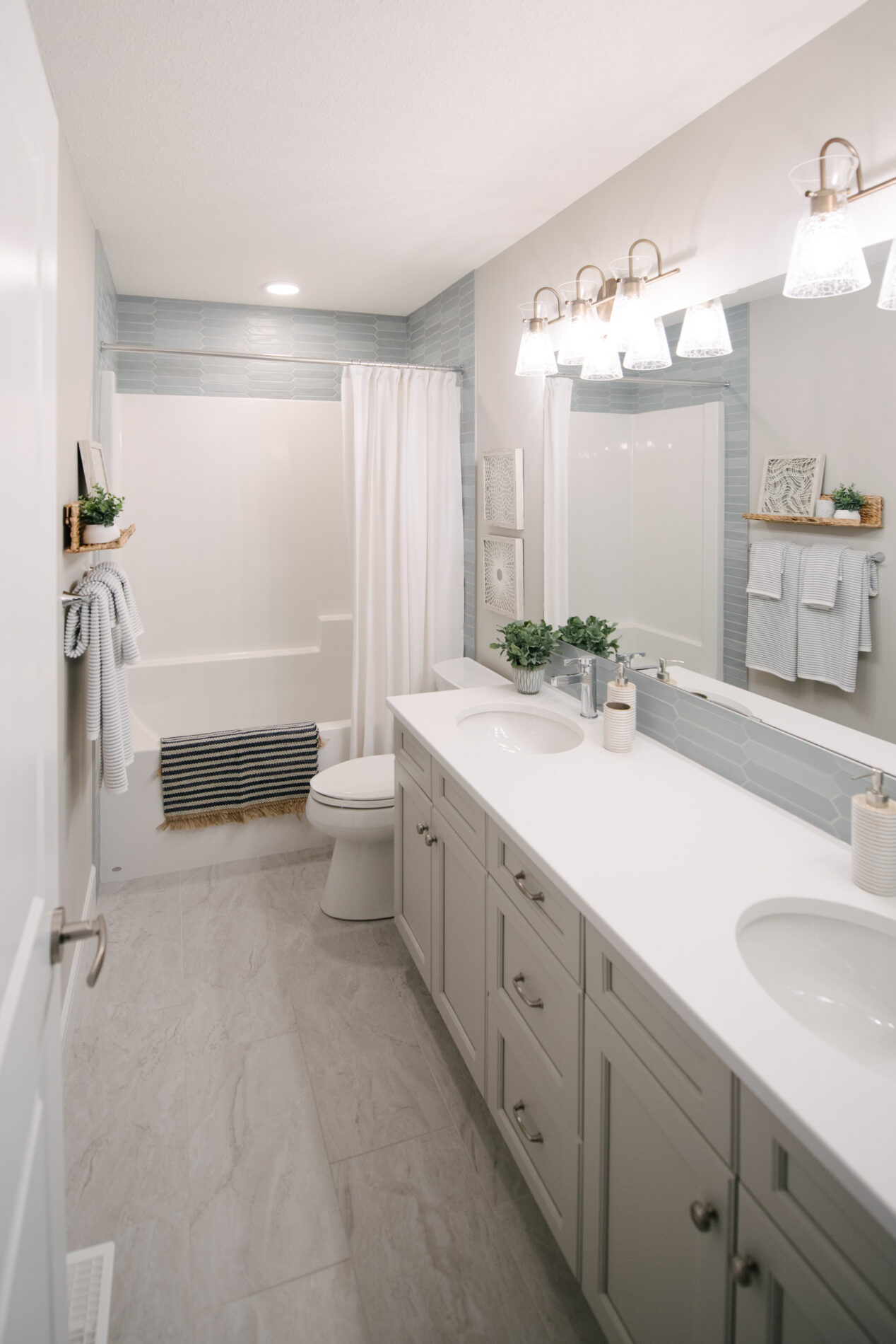 A large main bathroom with dual sinks, white countertops, warm grey/beige cabinets, and light blue backsplash tile. The light blue tile is carried around the tub/shower at the back of the room