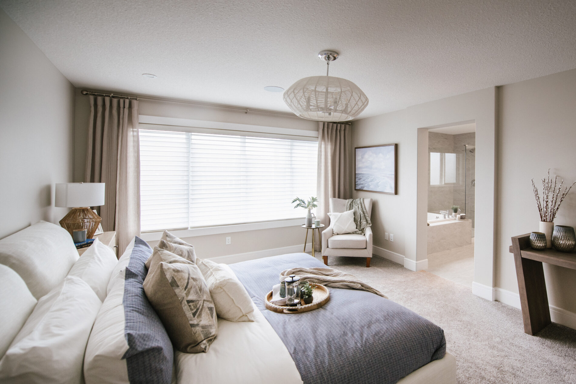 The calm, large master bedroom in the Kalliope showhome with warm grey bedding, a natural central light fixtures and a chair and small table in the corner