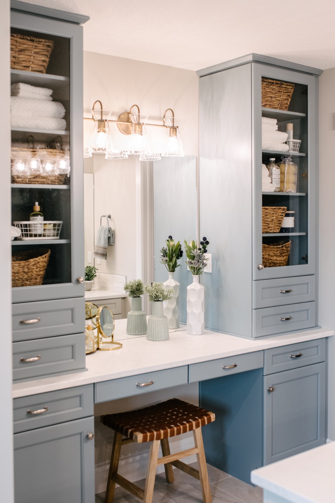 An oversized makeup desk in the Kalliope showhome ensuite with light blue cabinetry, two cabintery towers to either side of the desk with glass fronts showcasing organized bath accessories