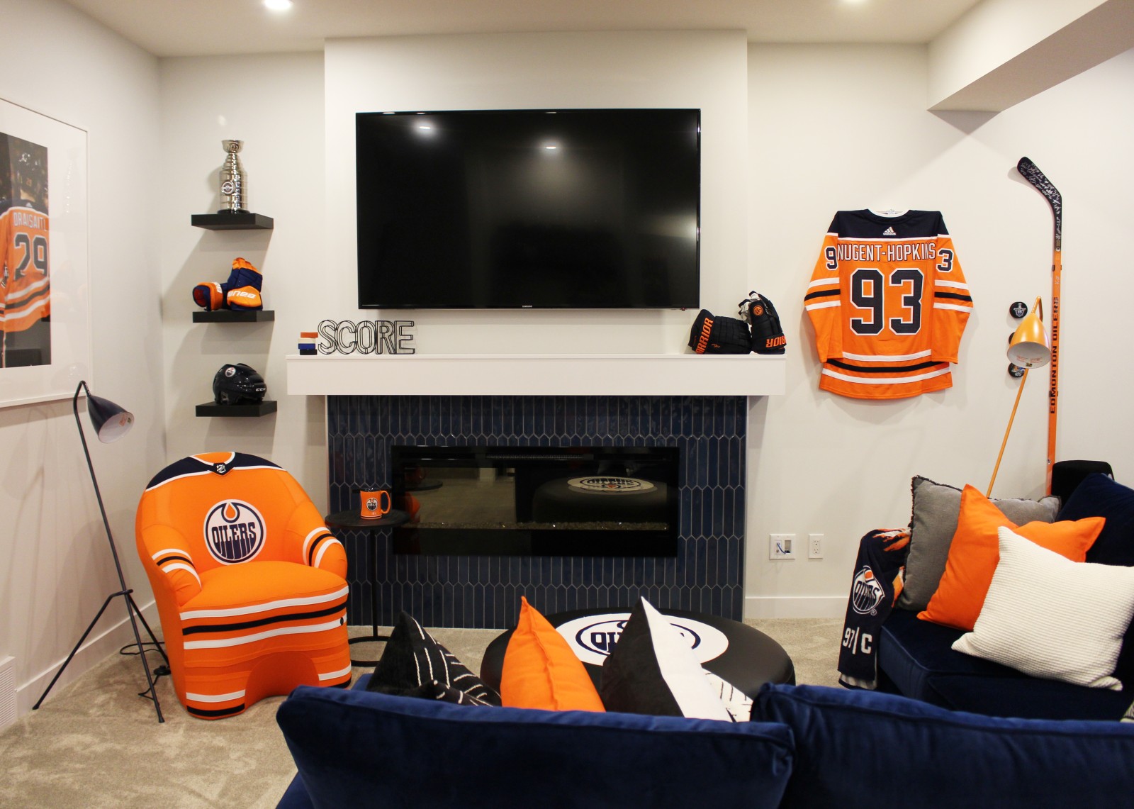 Oilers Fan Cave living room with blue accent tile on fireplace to match blue sectional couch, orange jersey chair, and hockey accessories