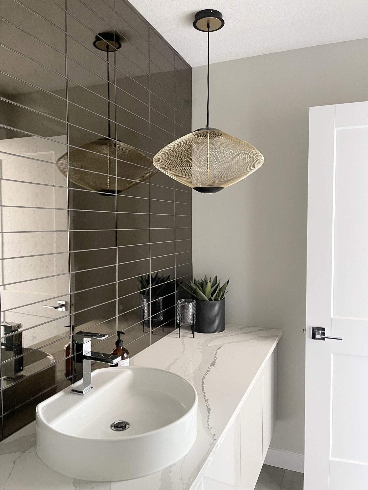 Modern gold light fixture hangs in front of a wall of mirrored tile in the half bath of the Cyrus showhome