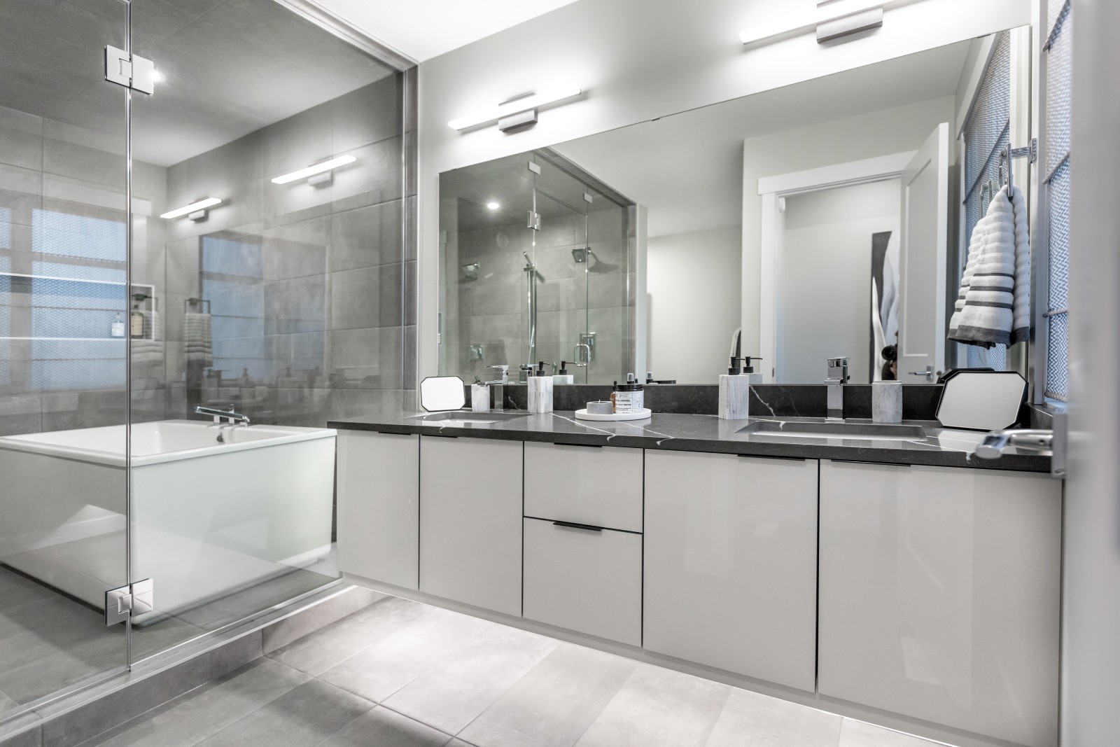 A floating vanity of grey acrylic cabinets with dark counter tops sits in front of the glass enclosed wet room
