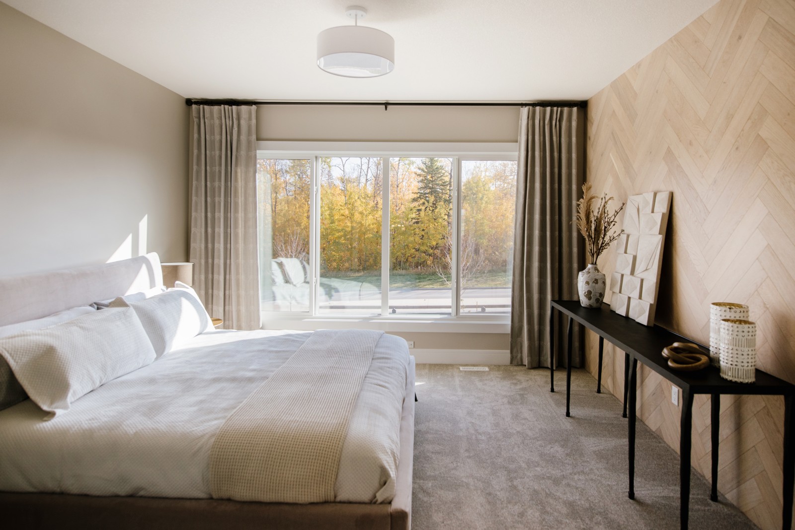 A warm master bedroom flooded with natural light from the wall of windows, highlighting the herringbone wood feature wall across from the bed