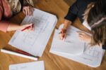 Overhead shot of two people looking and floorplans and marking them up with red pen